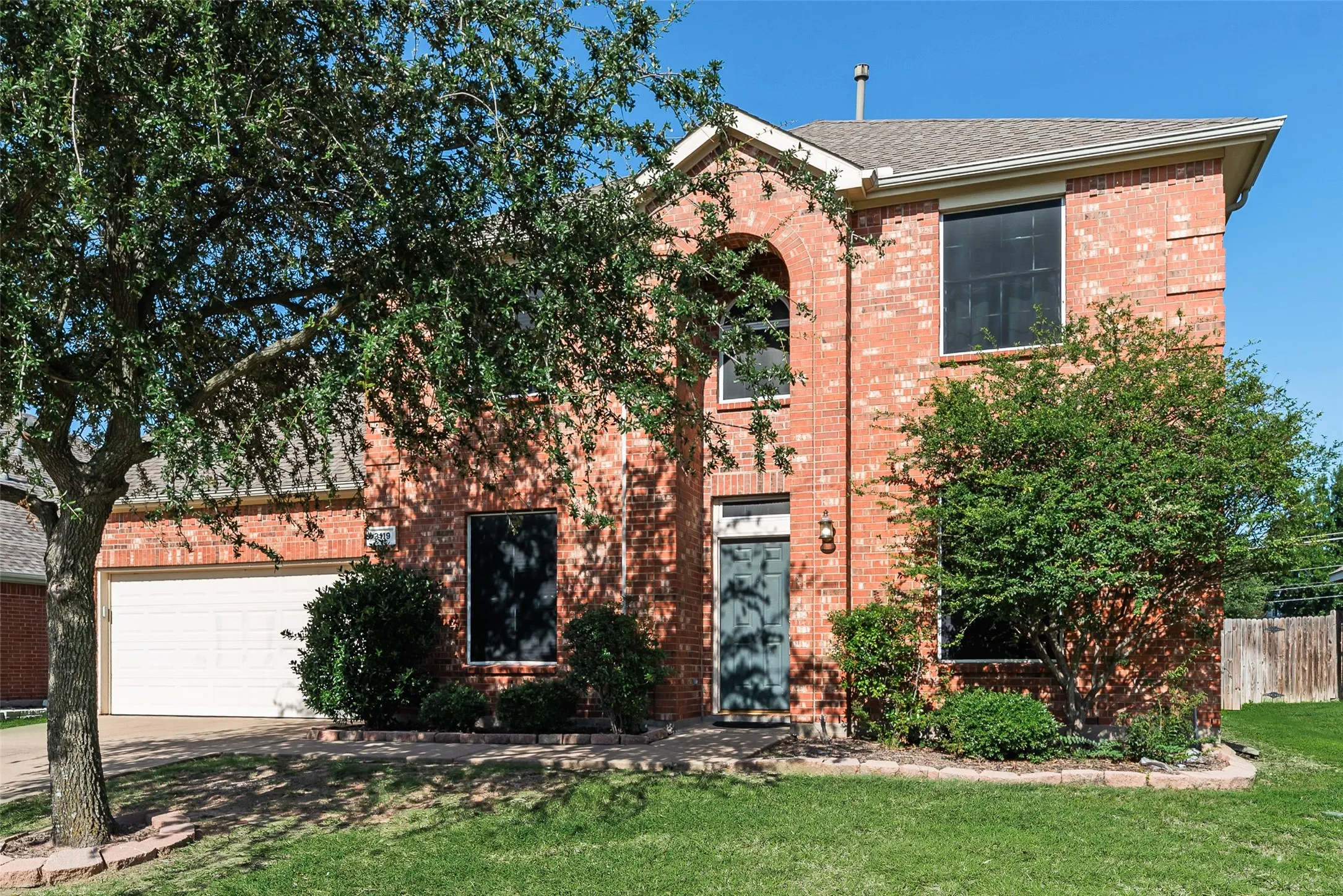 Charming two-story brick home featuring a white garage door, manicured lawn with mature shade trees, and ivy-covered exterior walls