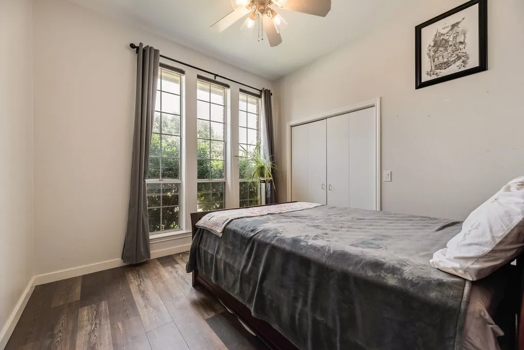Room featuring wood-finish flooring, three vertical windows with dark rods, a ceiling fan, and white bi-fold closet doors