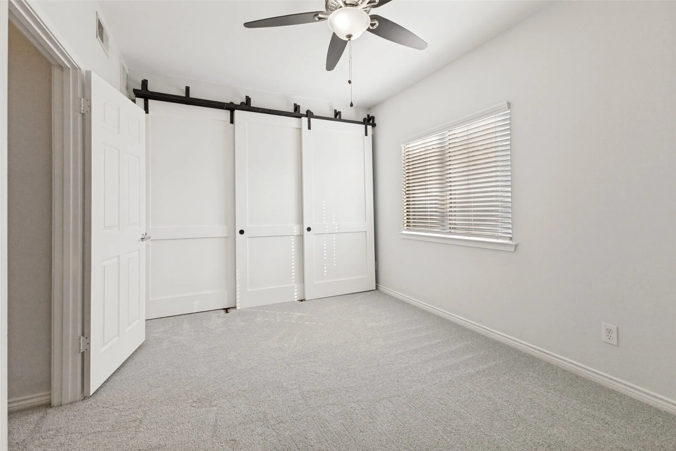 Spacious room featuring light gray carpeting, neutral wall paint, stately farm style doors for closet closure, and a window with horizontal blinds