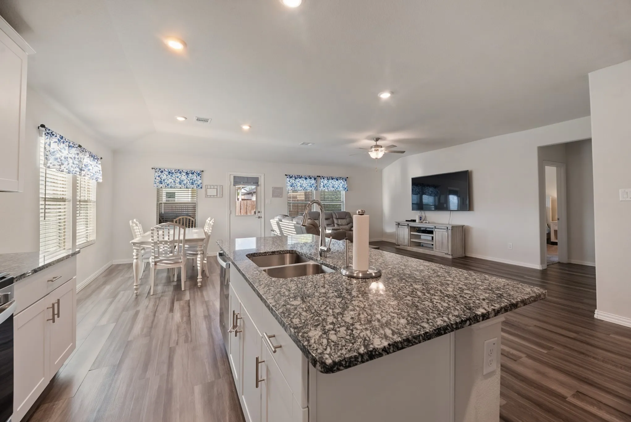 Spacious open-concept living area featuring a large kitchen island with a double basin sink and stone-finish countertop, white cabinetry with contemporary hardware, wood-finish flooring, and recessed lighting