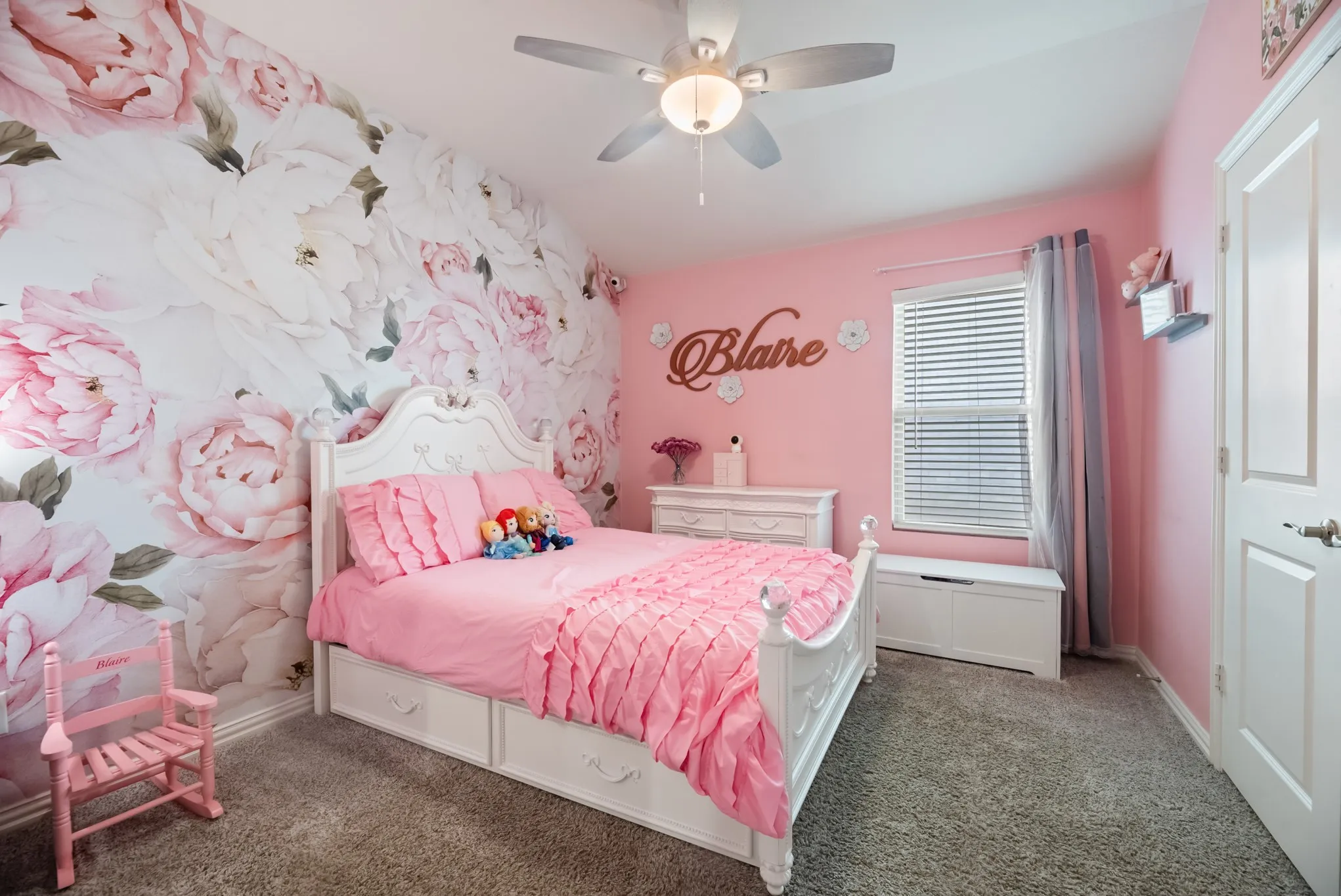 Bedroom featuring a floral accent wall, plush carpeting, ceiling fan with integrated lighting, a window with blinds, and an interior door with a brushed nickel handle