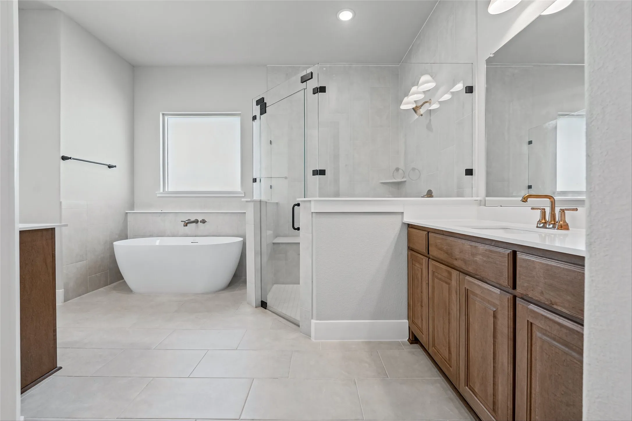 Freestanding soaking tub, glass-enclosed shower with black hardware, wood-finish vanity with white countertop, wall-mounted faucet, and light tile flooring
