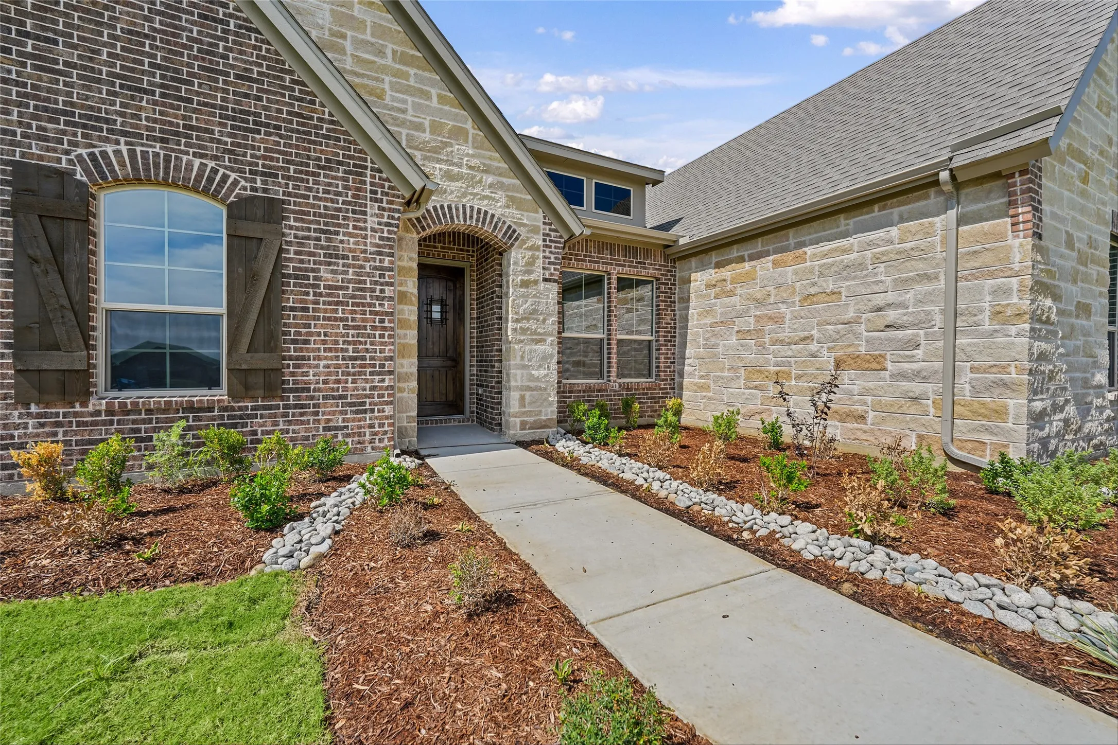 Brick and stone facade with arched entry, complementing wood shutters, and a concrete walkway bordered by landscaping