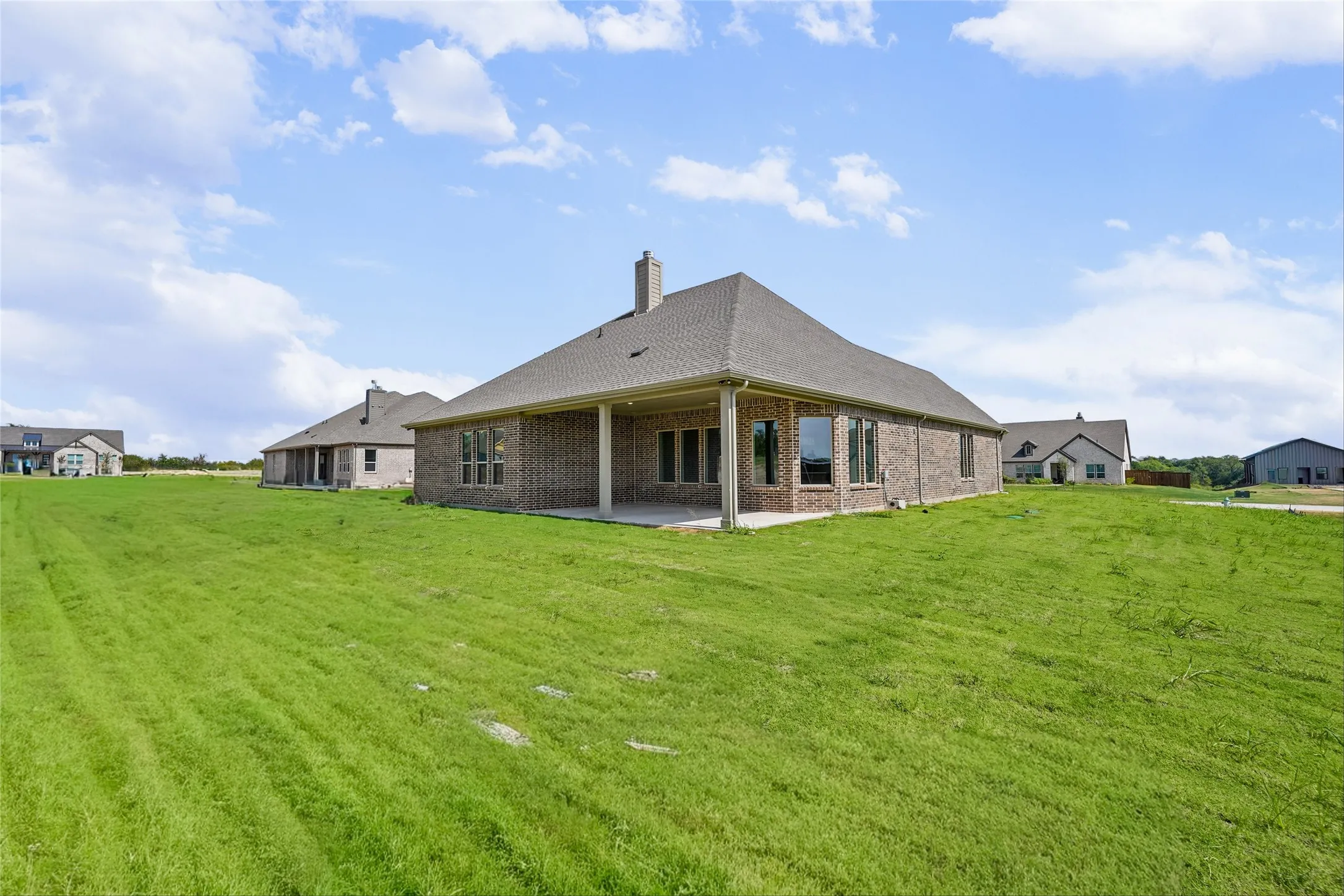 Expansive green space with a brick exterior, multiple windows, a covered patio, and a shingled roof