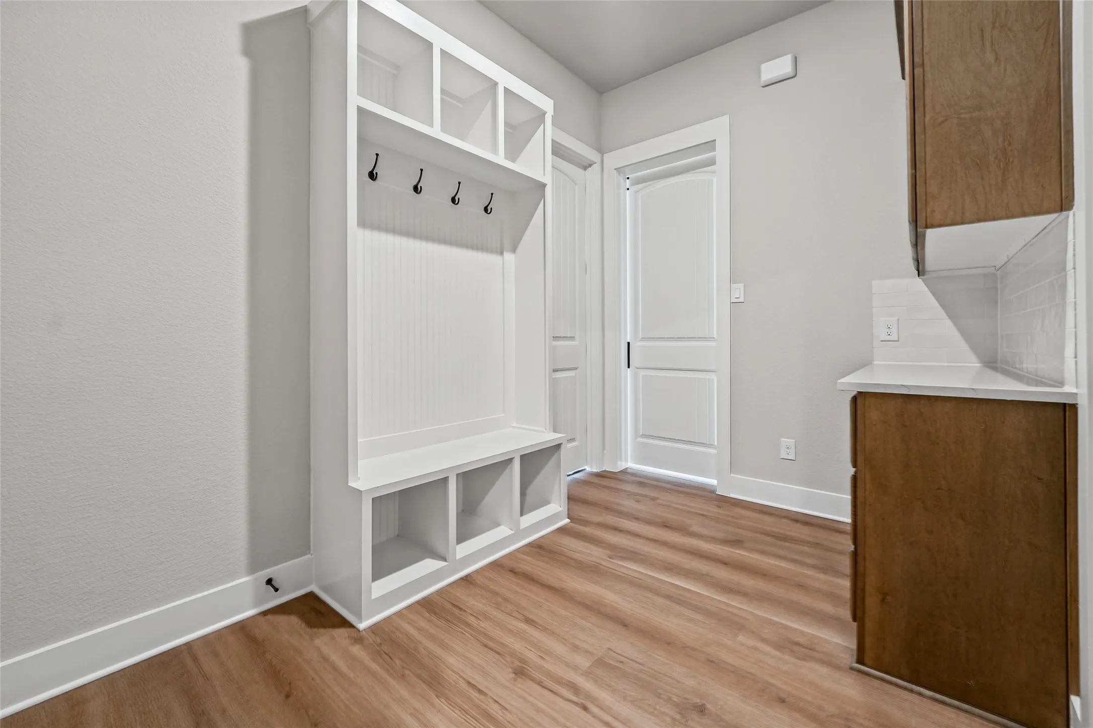 Built-in white storage unit with cubbies and coat hooks, wood-finish flooring, white doors with dark hardware, and wood-finish cabinetry with a white countertop and tile backsplash