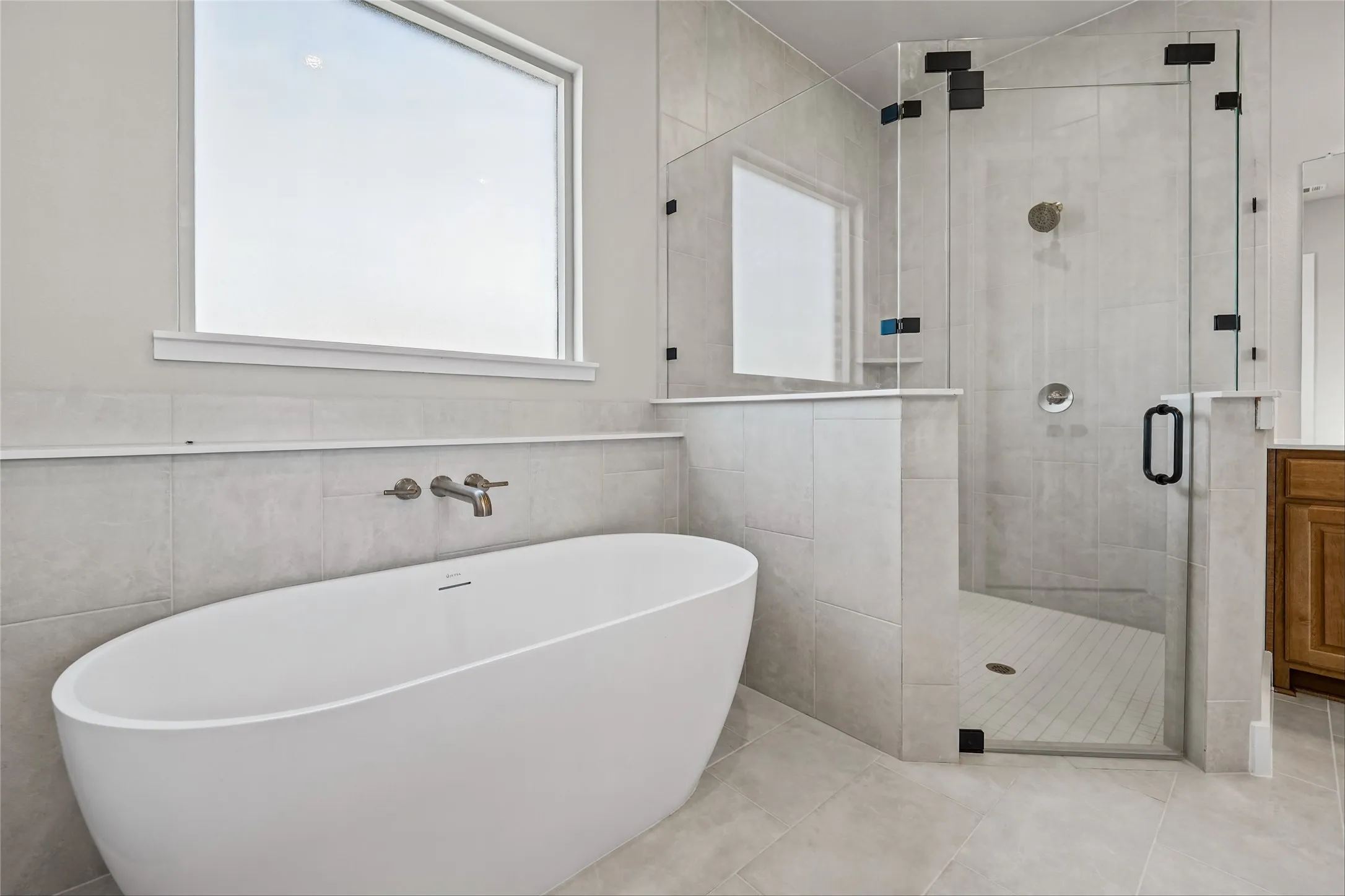 Freestanding soaking tub with wall-mounted bronze faucet, glass-enclosed shower with matte black hardware, and large format tile flooring