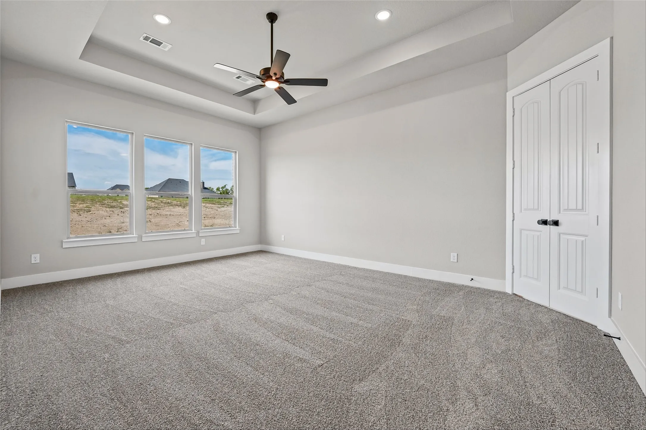 Tray ceiling with recessed lighting and a ceiling fan, three large windows, gray carpeting, and a double-door entry