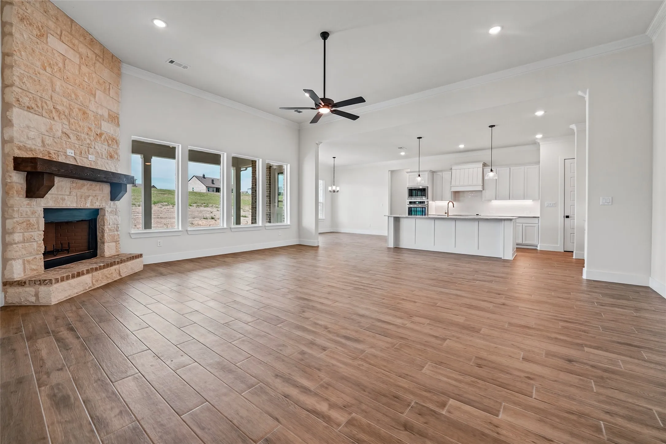 Spacious great room with wood-finish flooring, stone-clad fireplace with wood mantel, and a wall of windows