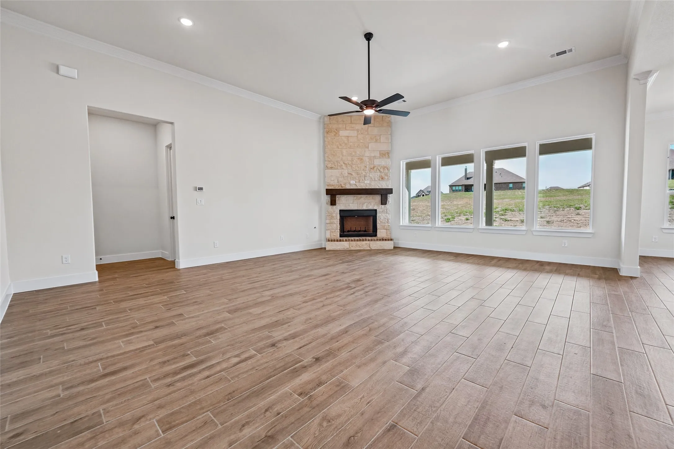 Spacious living area featuring wood-finish flooring, a stone fireplace with a wood mantle, a ceiling fan, and multiple large windows