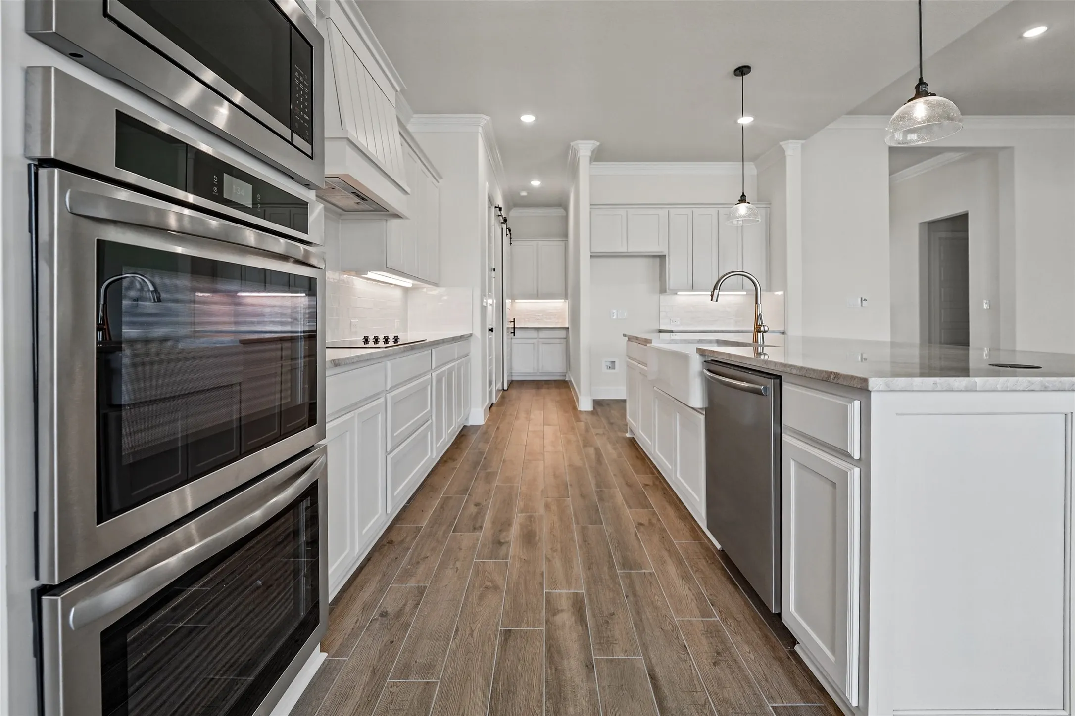 Gourmet kitchen featuring wood-finish flooring, white cabinetry, stainless steel double ovens, a farmhouse sink, and a center island with pendant lighting
