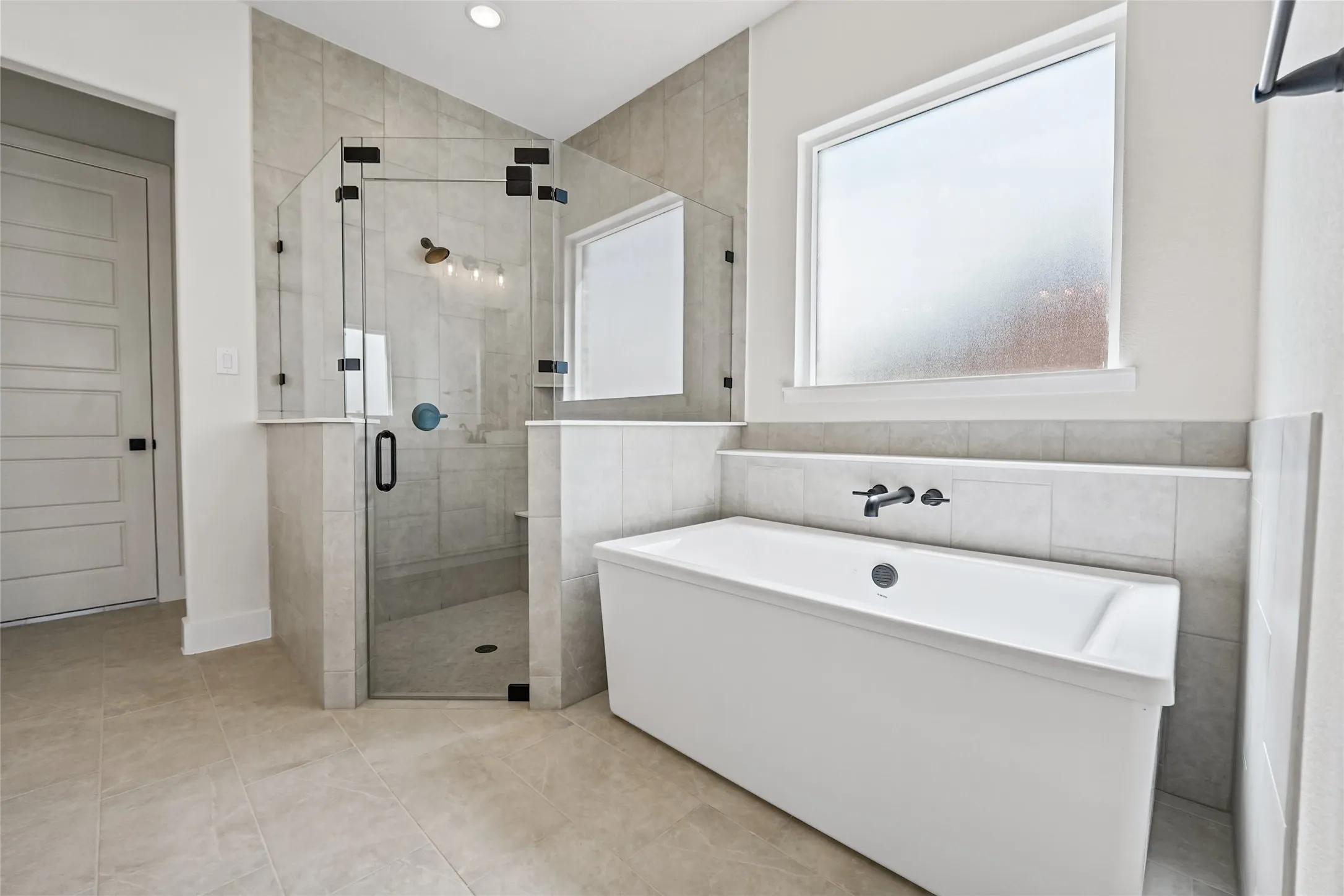 Tiled flooring, freestanding soaking tub with wall-mounted faucet, frameless glass shower enclosure, and frosted window