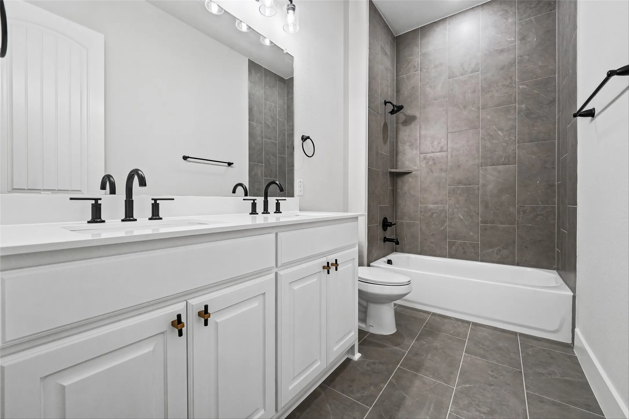 Dual vanity with white cabinetry and matte black faucets, complemented by a white bathtub with a tile surround, and gray tile flooring