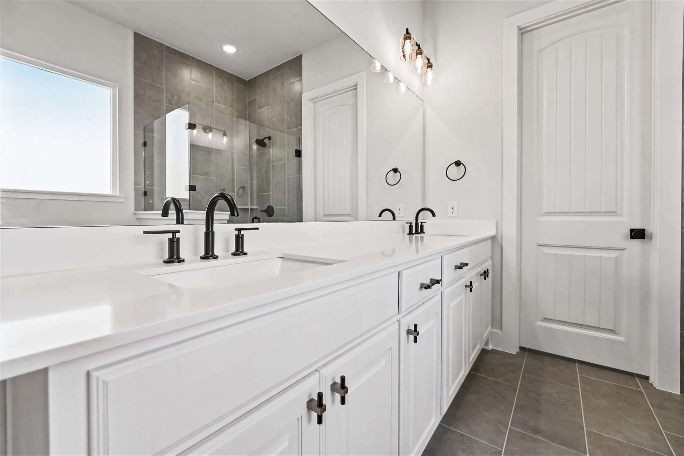 White dual vanity with integrated sinks, matte black hardware, large wall mirror, and a window providing natural light