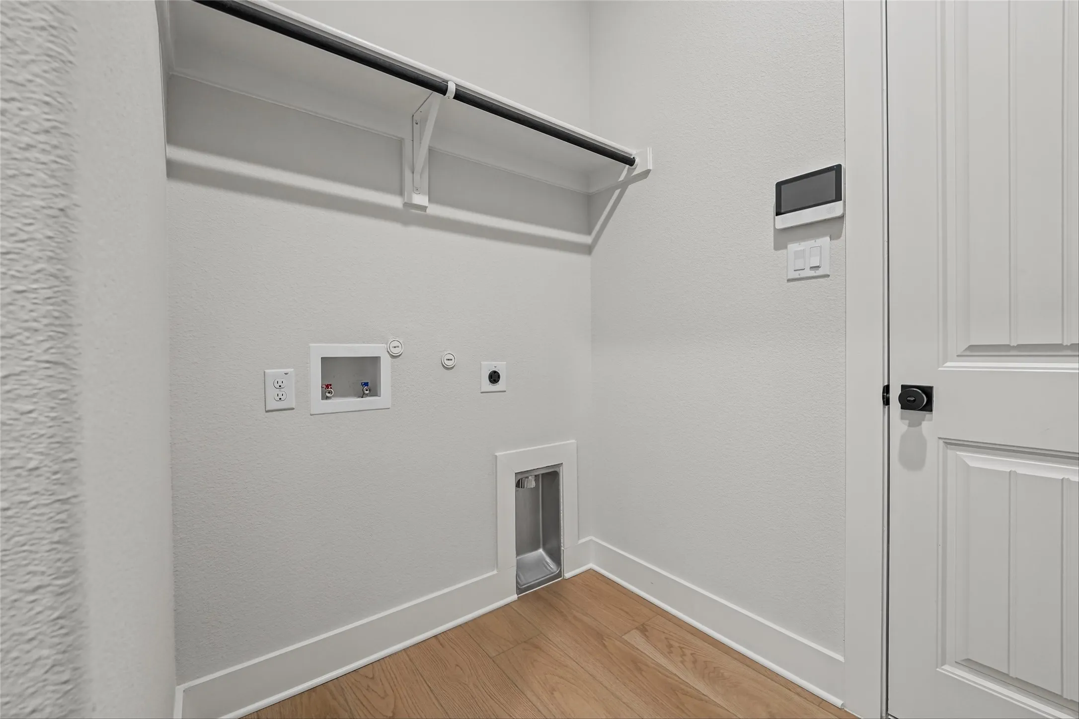 Bright laundry area featuring wood-finish flooring, a wall-mounted shelf with a hanging rod, and a panel door with black hardware