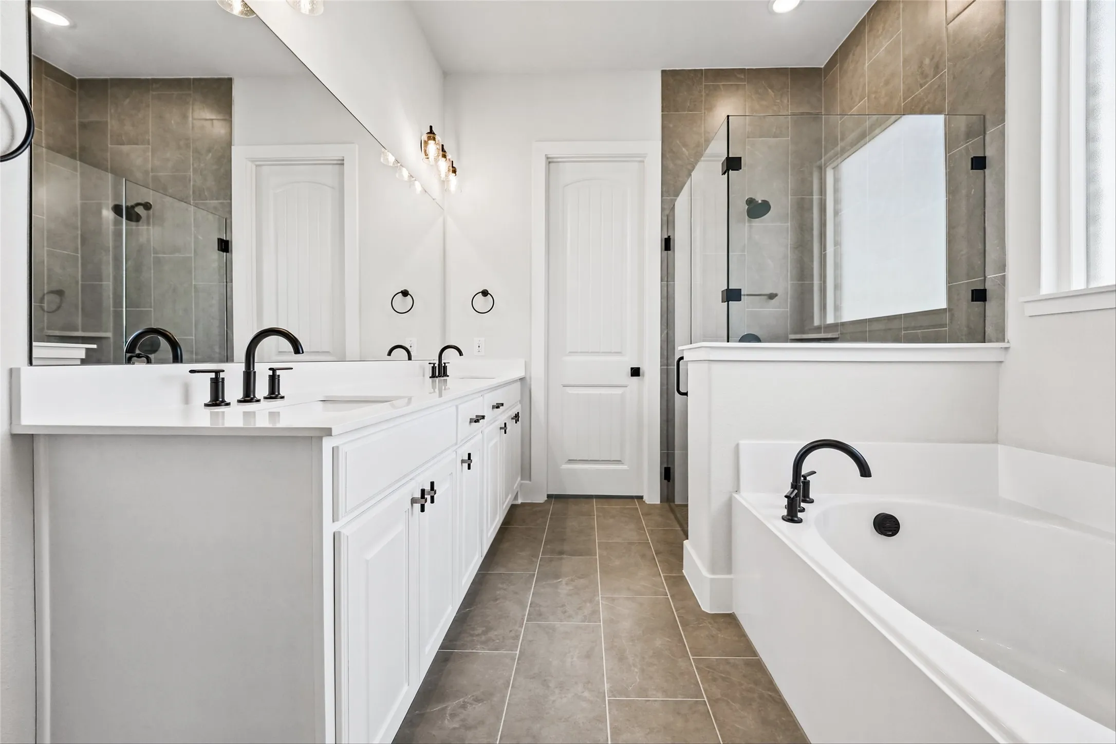 Dual vanity with white cabinetry, matte black fixtures, an expansive mirror, a glass-enclosed shower, and a built-in soaking tub