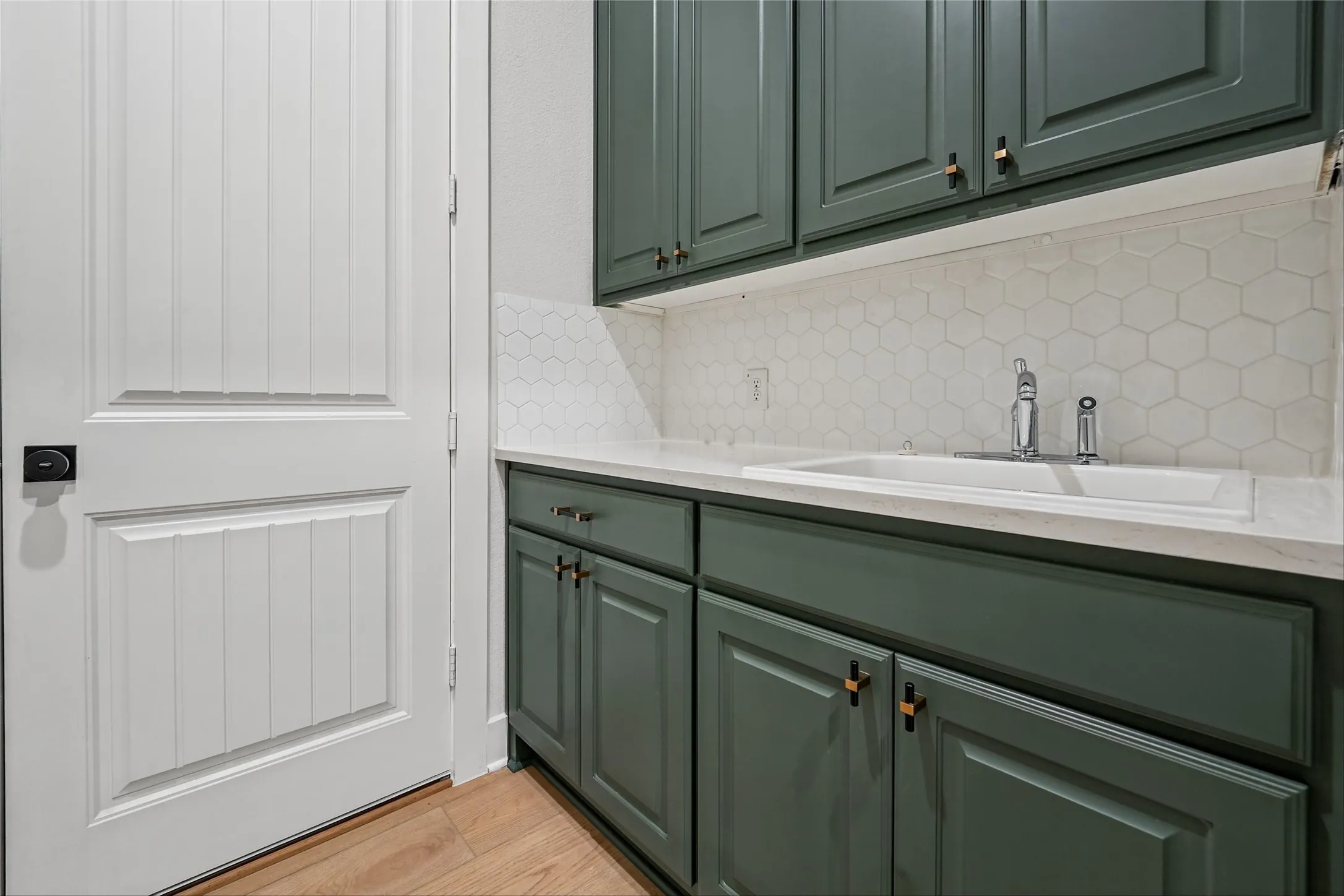Green shaker-style cabinetry with gold-tone hardware, white hexagon tile backsplash, integrated sink, light-toned countertop, and wood-finish flooring