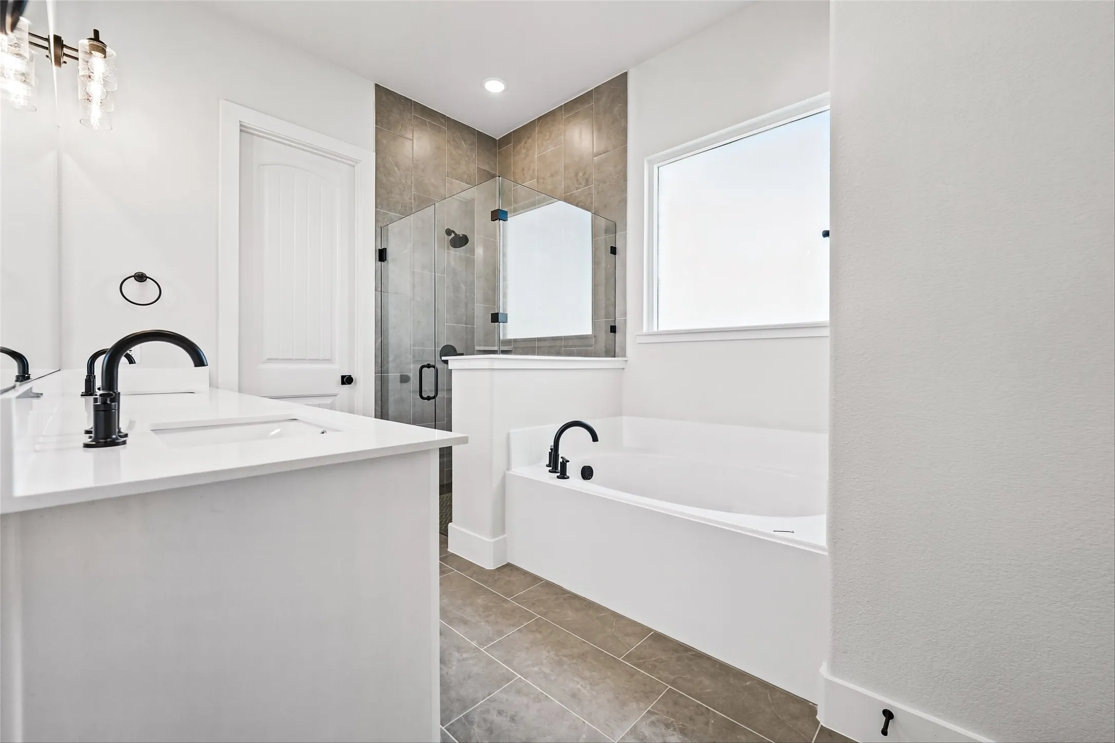 Dual vanity with integrated sinks and matte black fixtures, soaking tub with matte black fixtures, enclosed shower with glass surround and matte black hardware, tiled shower surround, and tiled flooring