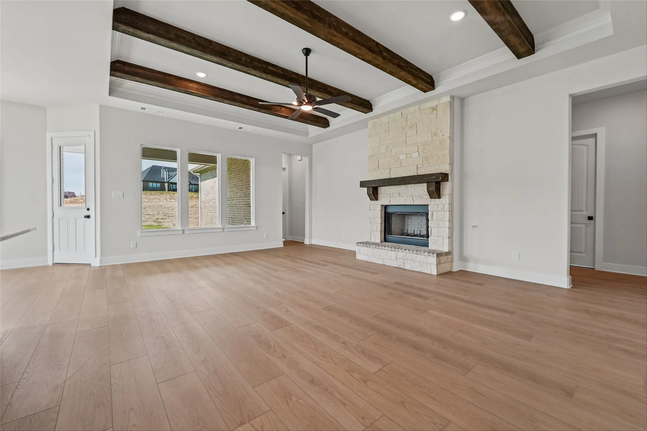 Spacious room featuring wood-finish flooring, a stone-clad fireplace with a wood mantel, exposed ceiling beams, and a ceiling fan