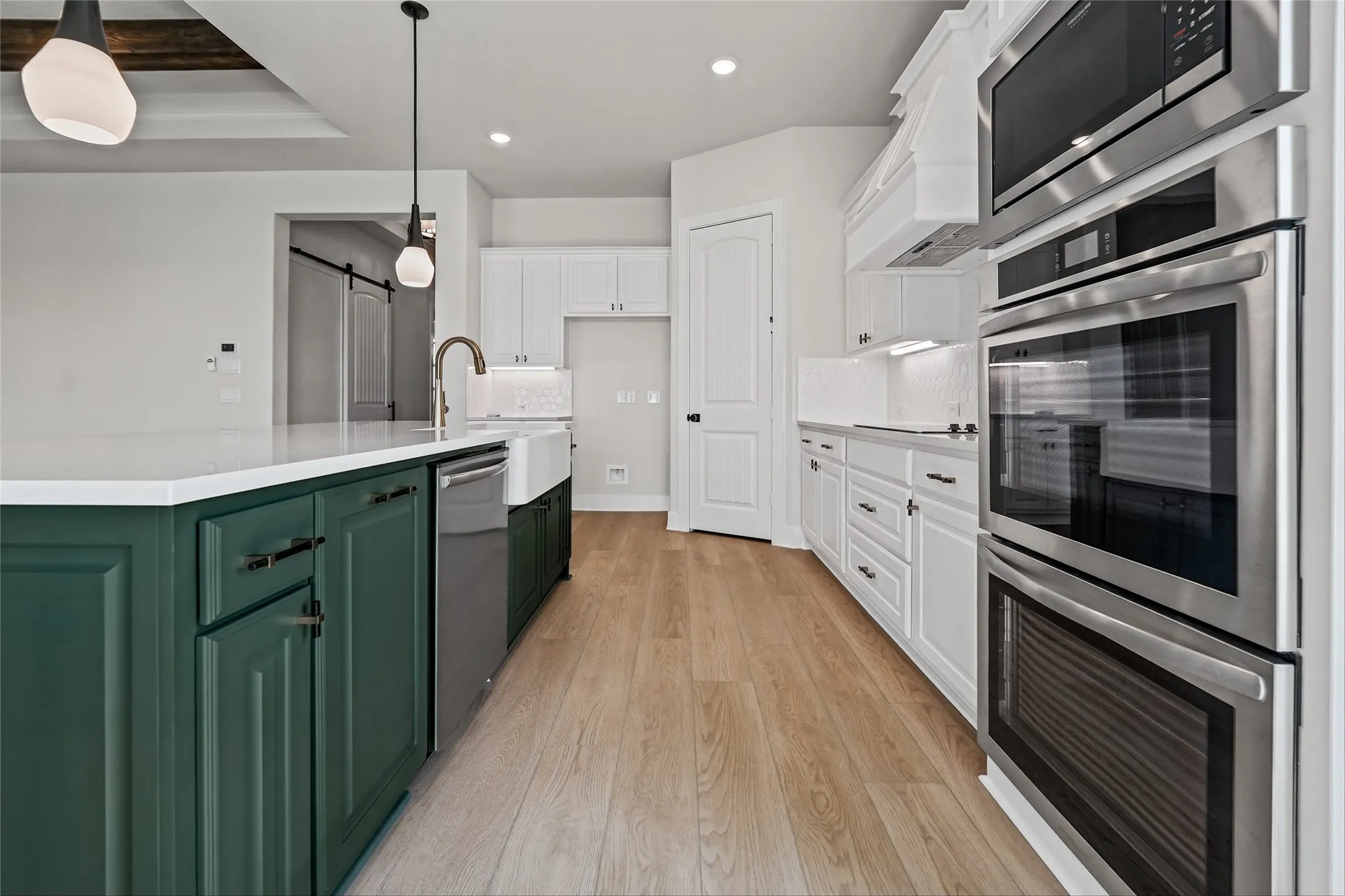 Gourmet kitchen with white cabinetry and dark green island, stainless steel built-in appliances, farmhouse sink with a gooseneck faucet, light wood-finish flooring, and recessed lighting