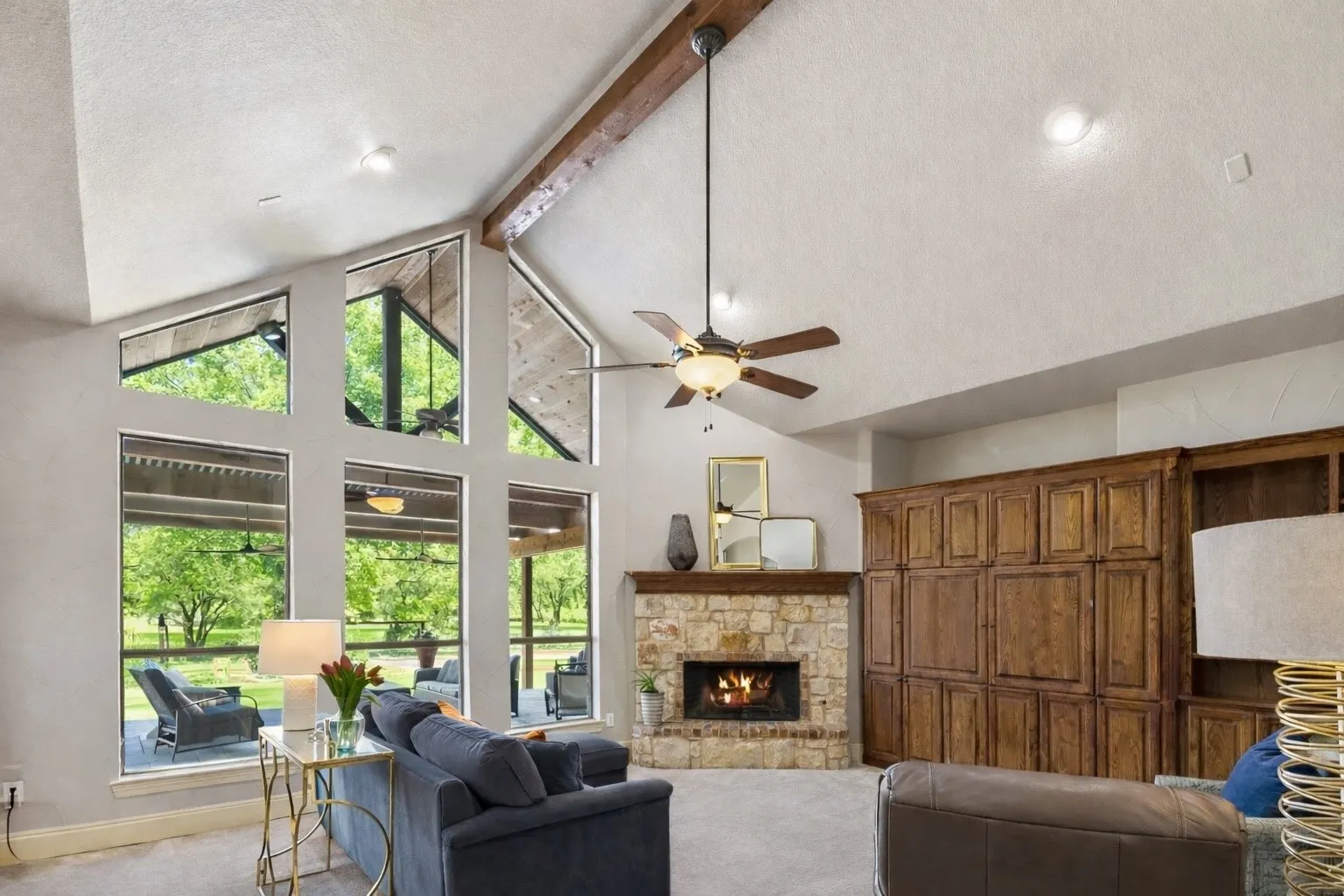 The spacious living room features floor-to-ceiling windows that flood the space with natural light while overlooking the wooded backyard, a gorgeous stone fireplace, stained wood built-ins, and a vaulted ceiling with an exposed beam.