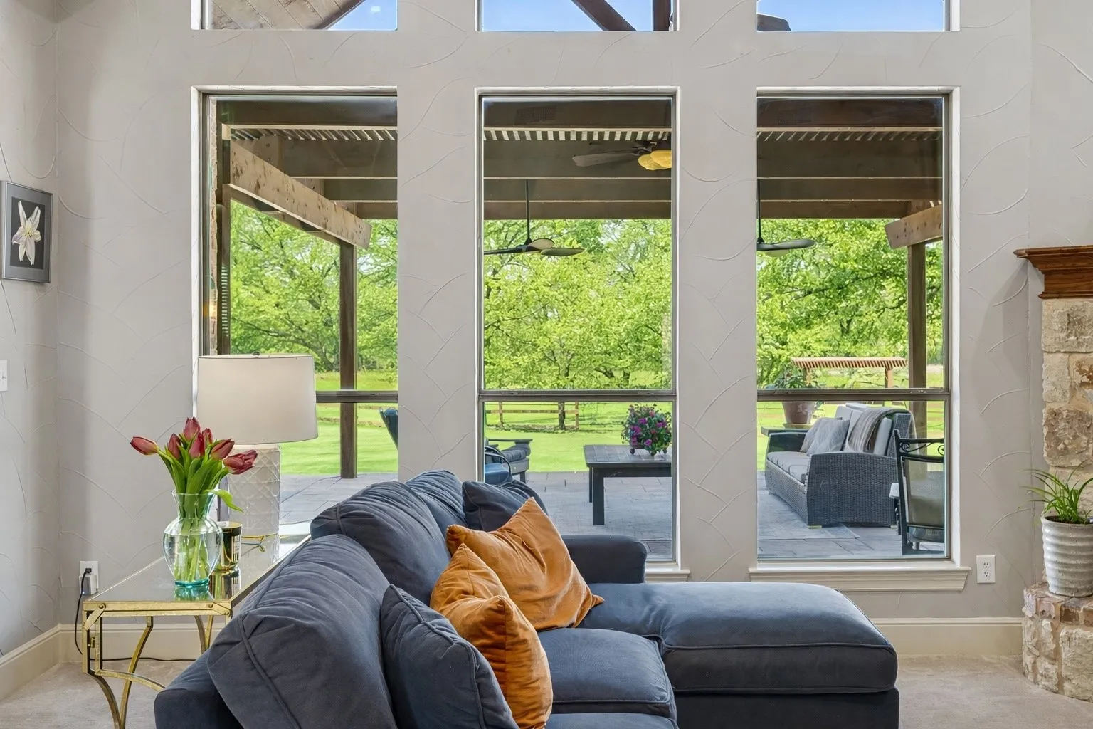 View of the amazing backyard through the tall living room windows. Imagine enjoying this view overlooking your outdoor patio and verdant landscape every day! This warm and welcoming living area is centered around the expansive outdoor views and the beautiful stone fireplace complemented by custom built-ins.