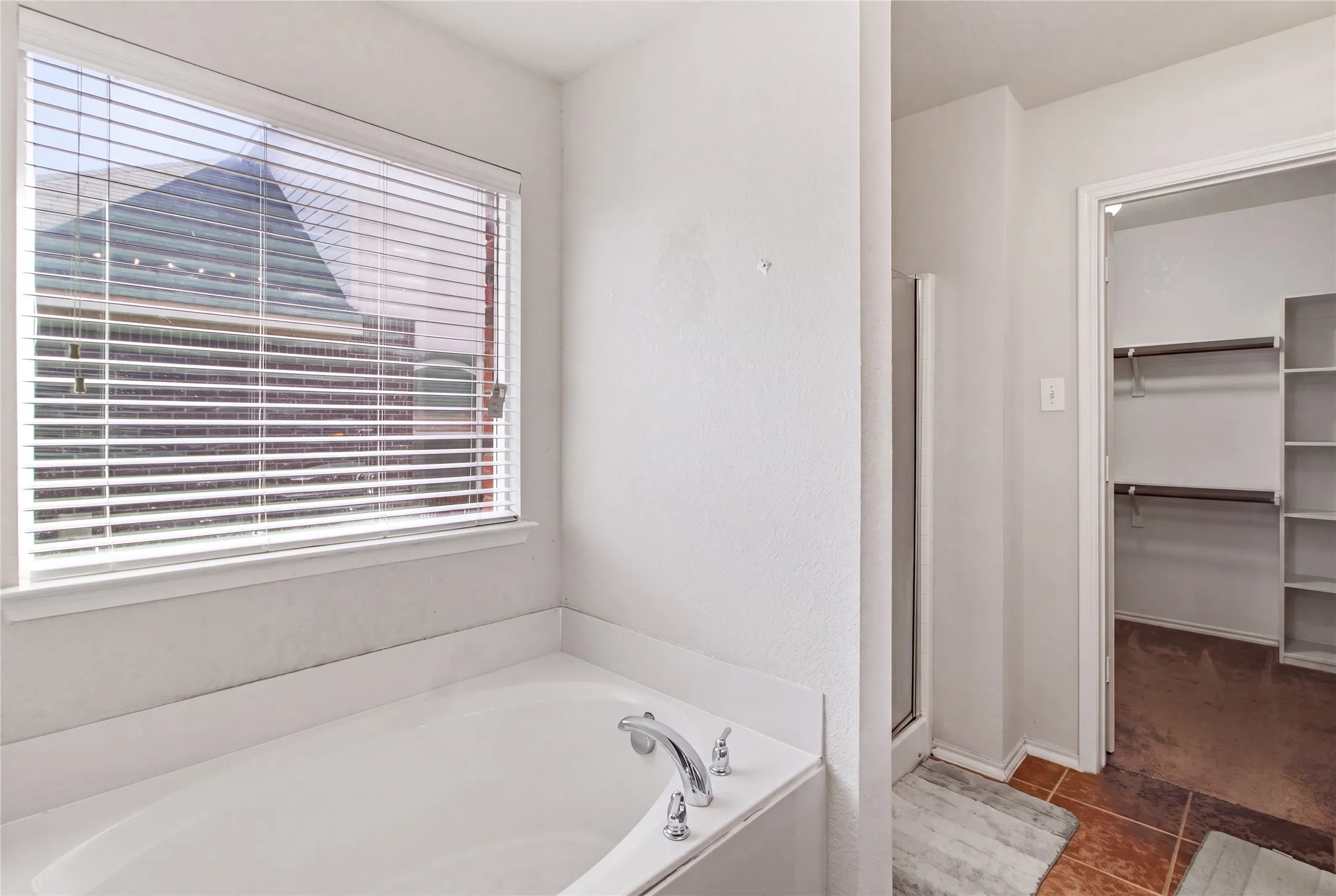 Built-in soaking tub with chrome fixtures, large window with blinds, adjacent walk-in shower with glass enclosure, and a nearby closet with shelving and hanging rods