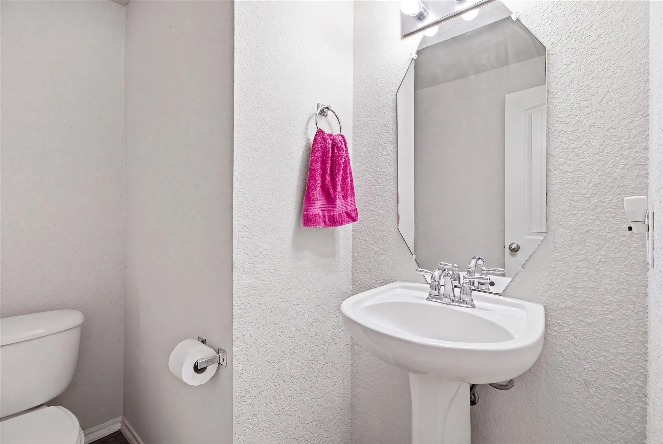 Pedestal sink featuring a chrome faucet, illuminated by a vanity light fixture, with a geometric mirror