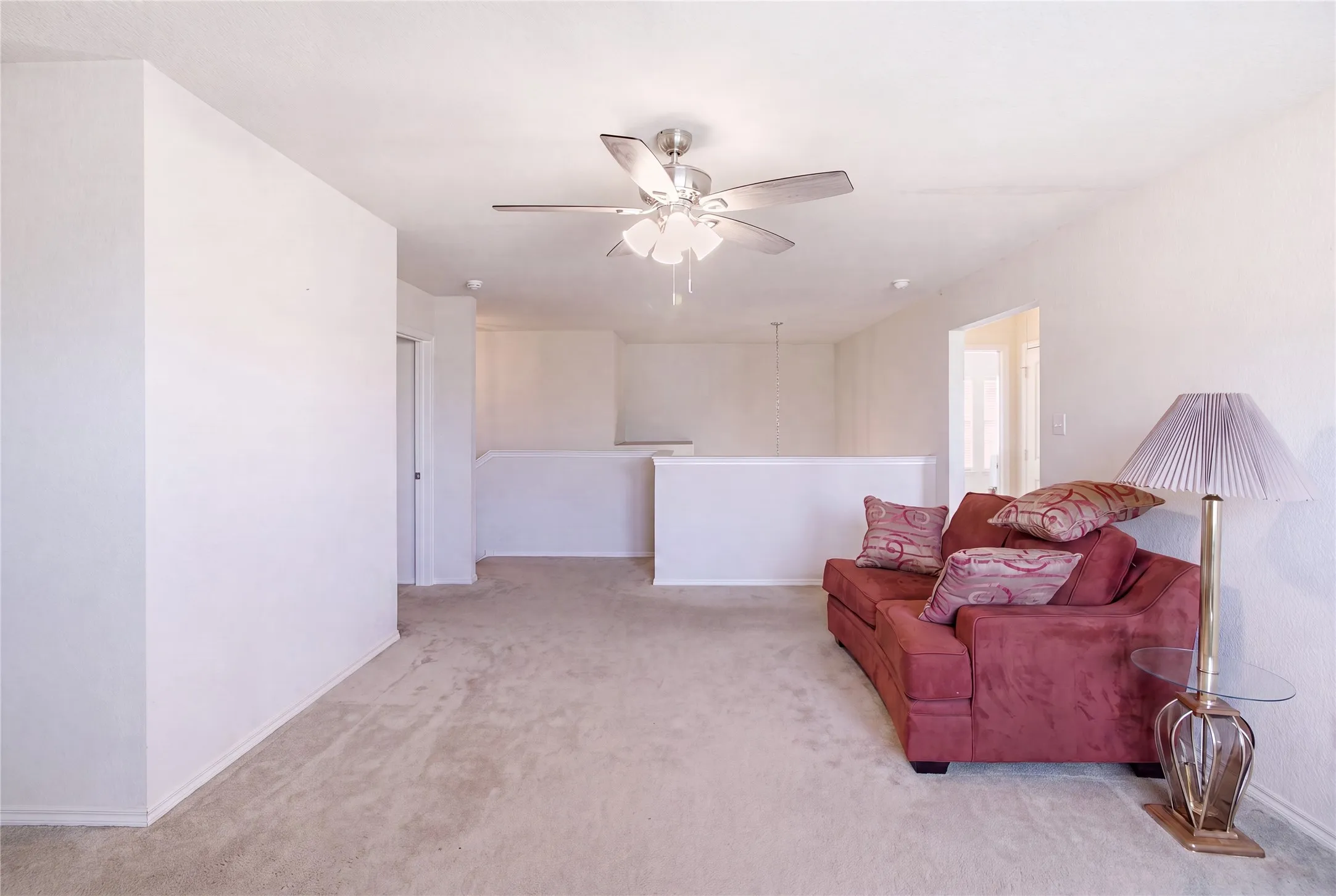 Spacious carpeted room featuring a ceiling fan with integrated lighting, neutral wall tones, and a partial wall overlooking an open area