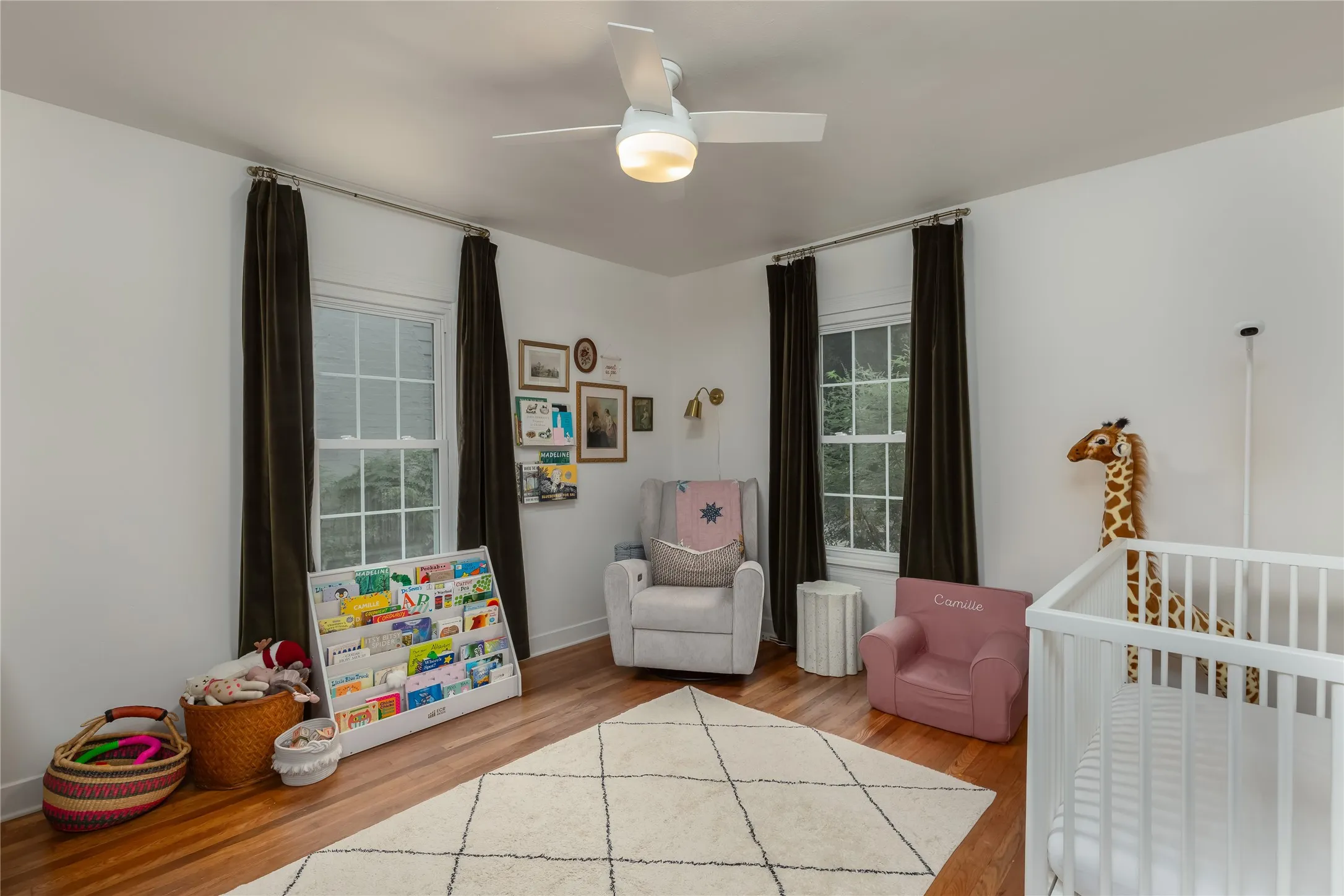 Bright room featuring wood-finish flooring, two large windows with dark drapery, a ceiling fan with integrated lighting, and a wall-mounted sconce