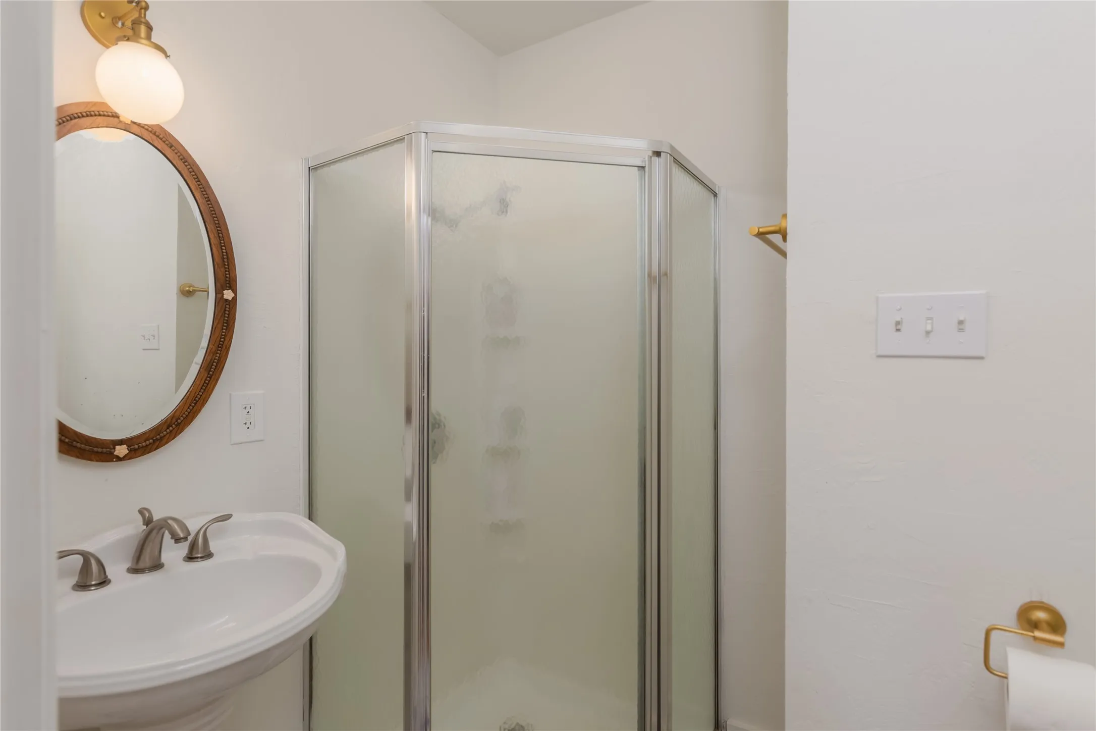 Enclosed corner shower with frosted glass panels, pedestal sink with brushed nickel faucet, and an oval mirror with a decorative wood frame