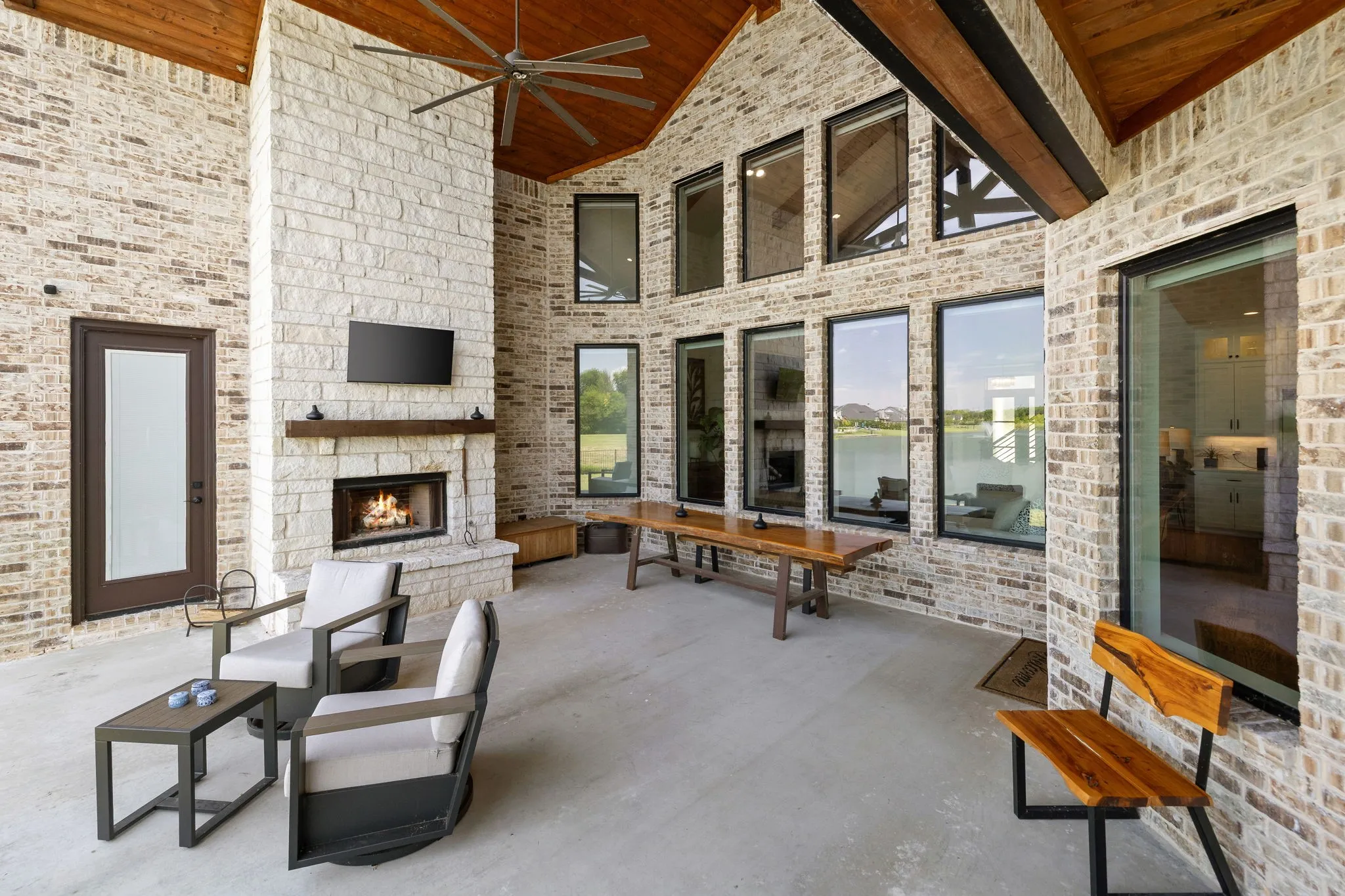 Expansive covered patio featuring a full outdoor kitchen, a stone fireplace, brick exterior walls, multiple floor-to-ceiling windows, and a wood-paneled ceiling