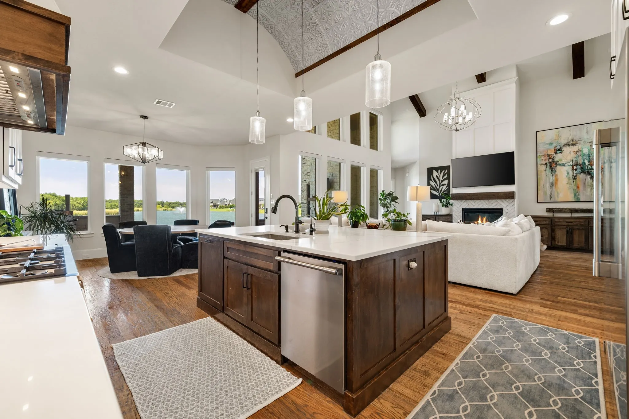 Open-concept living space featuring wood-finish flooring, a kitchen island with stone countertops and integrated appliances, a vaulted ceiling with exposed beams, a fireplace with a stone surround, and large windows overlooking a body of water