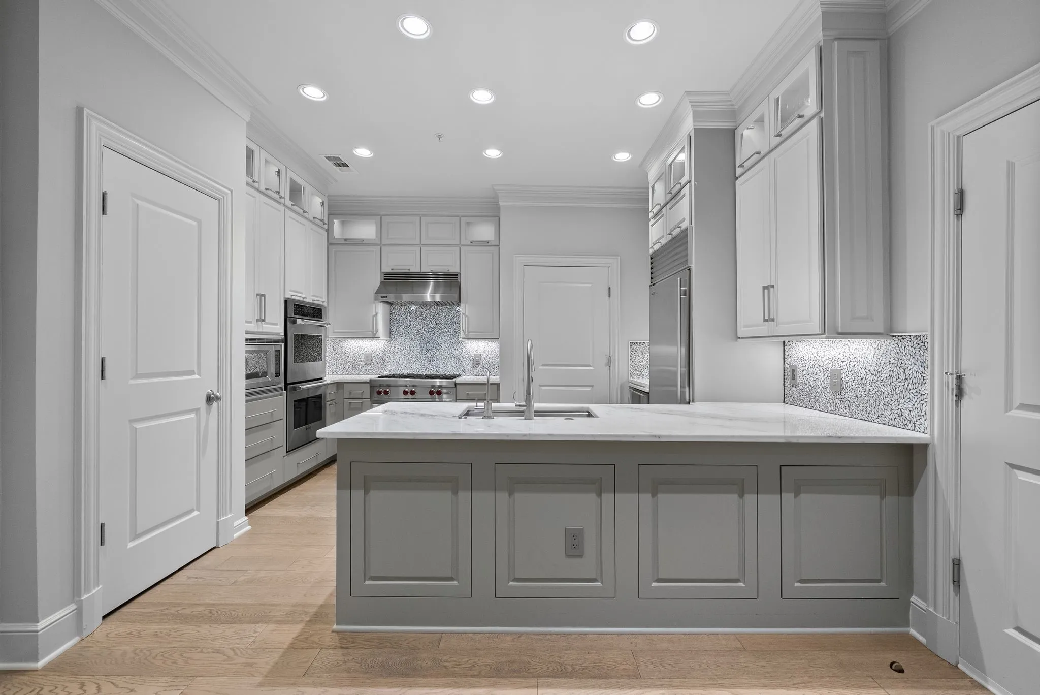 Gourmet kitchen featuring a central island with a sink, light wood-finish flooring, recessed lighting, and stainless steel appliances, including a range hood and double ovens