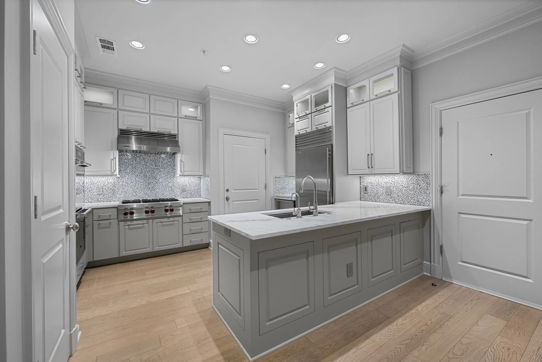 Gourmet kitchen featuring a large central island with an undermount sink, a professional-grade gas range with stainless steel hood, custom cabinetry with glass-front uppers, a mosaic tile backsplash, and wood-finish flooring