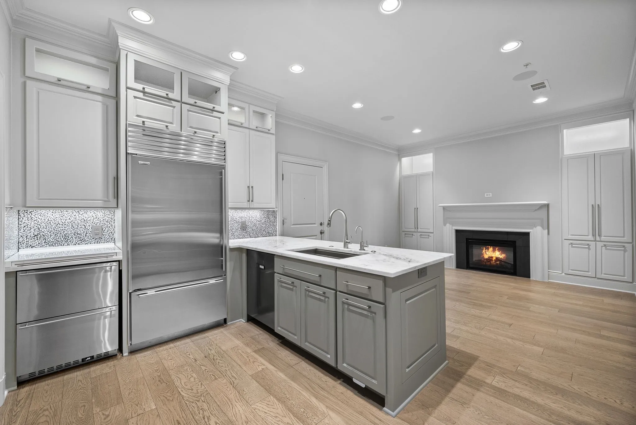 Modern kitchen featuring a central island with an undermount sink, stainless steel appliances, light wood-finish flooring, recessed lighting, and a fireplace with a white mantel