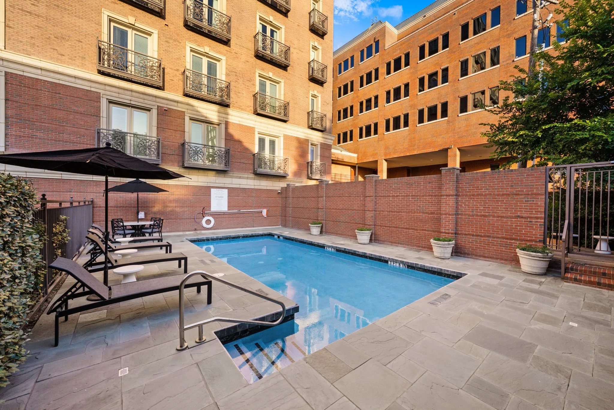 Outdoor swimming pool area featuring a perimeter of large format paver stones, a brick privacy wall, and a stainless steel pool railing