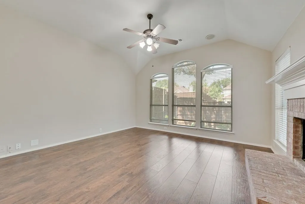 Vaulted ceiling room featuring wood-finish flooring, three arched windows with blinds, a ceiling fan, and a brick fireplace with a white mantel