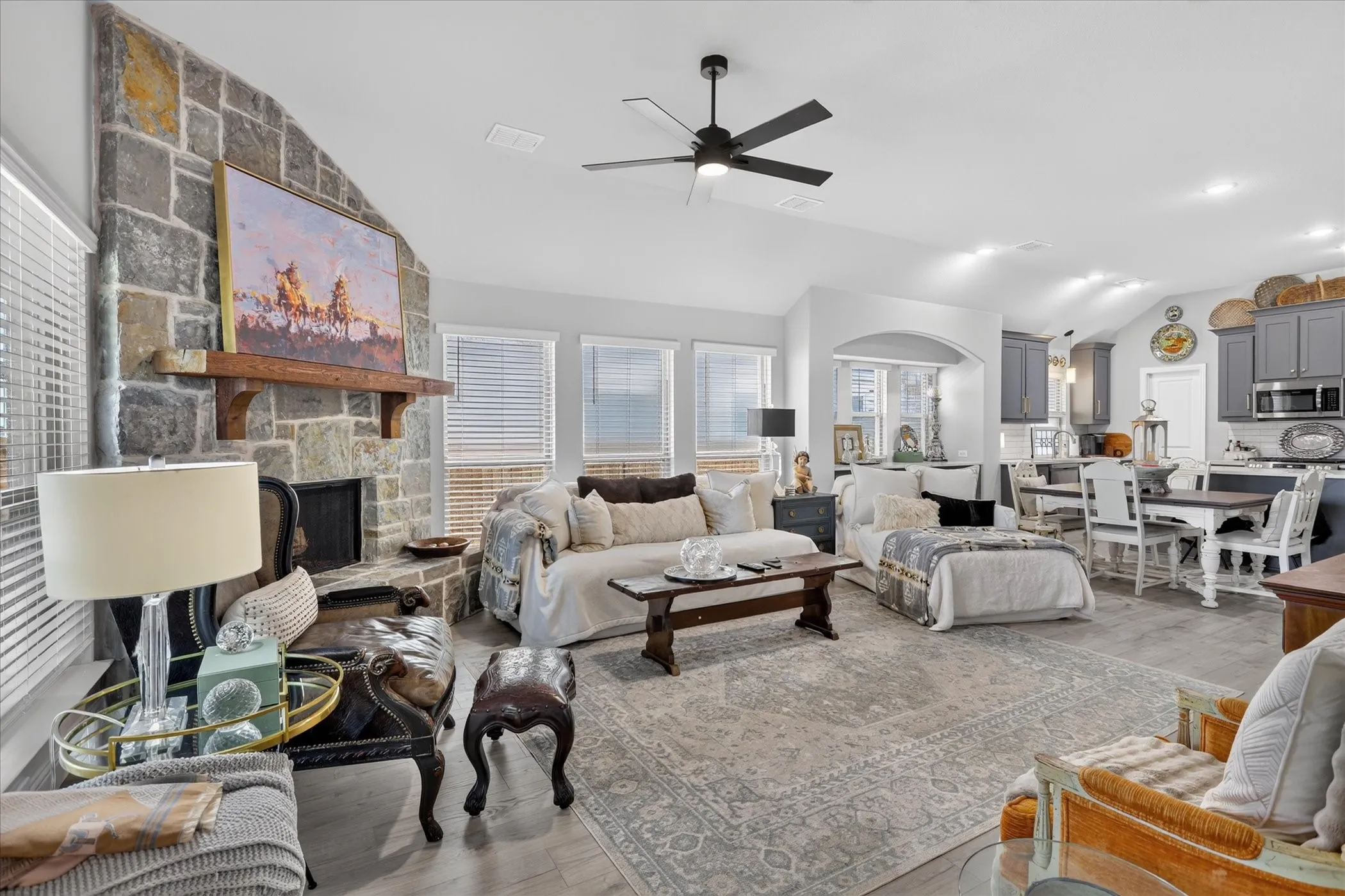 Expansive living room featuring a stone fireplace with a rustic wood mantel, multiple windows with horizontal blinds, and wood-finish flooring