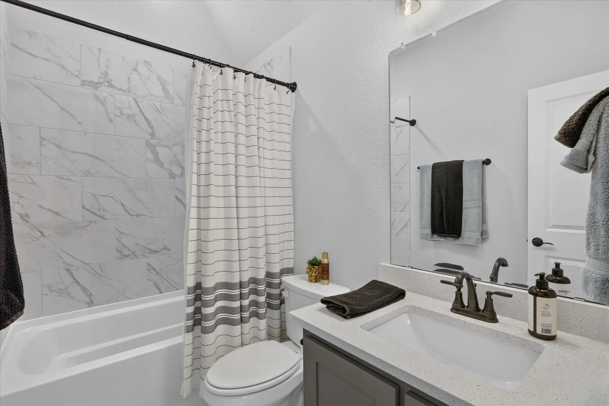 Bathroom featuring a white bathtub, large format marble-look tile surround, dark gray vanity with a light speckled countertop, undermount sink, and matte bronze fixtures