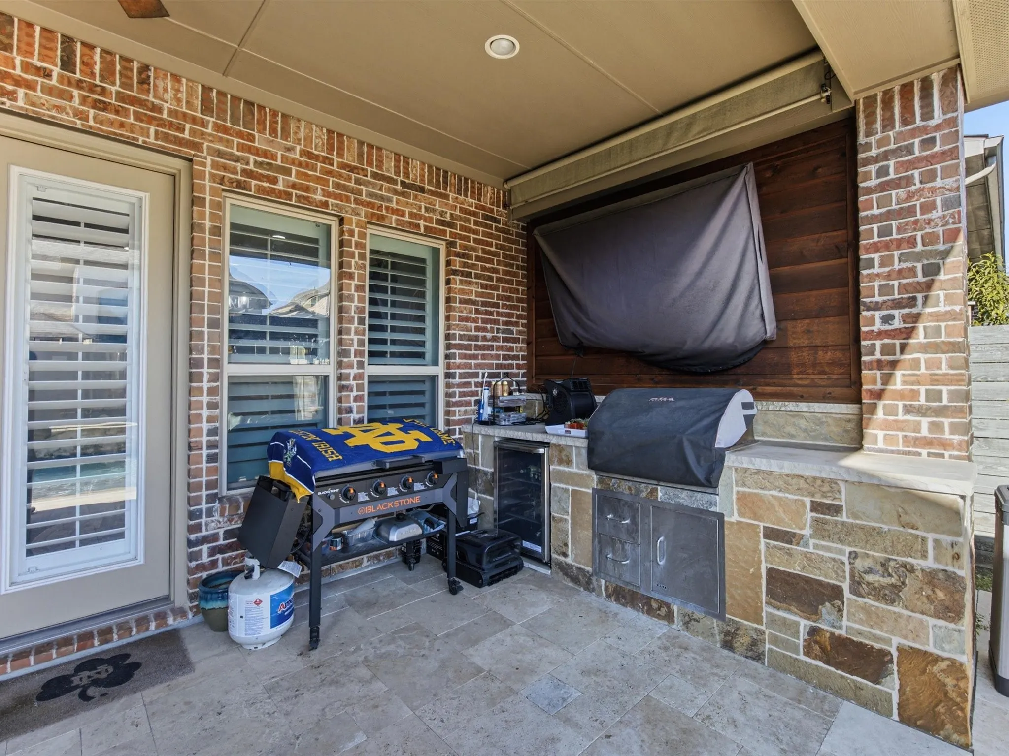 Covered outdoor kitchen featuring a built-in grill, integrated beverage refrigerator, stone-clad island, and wood accent wall