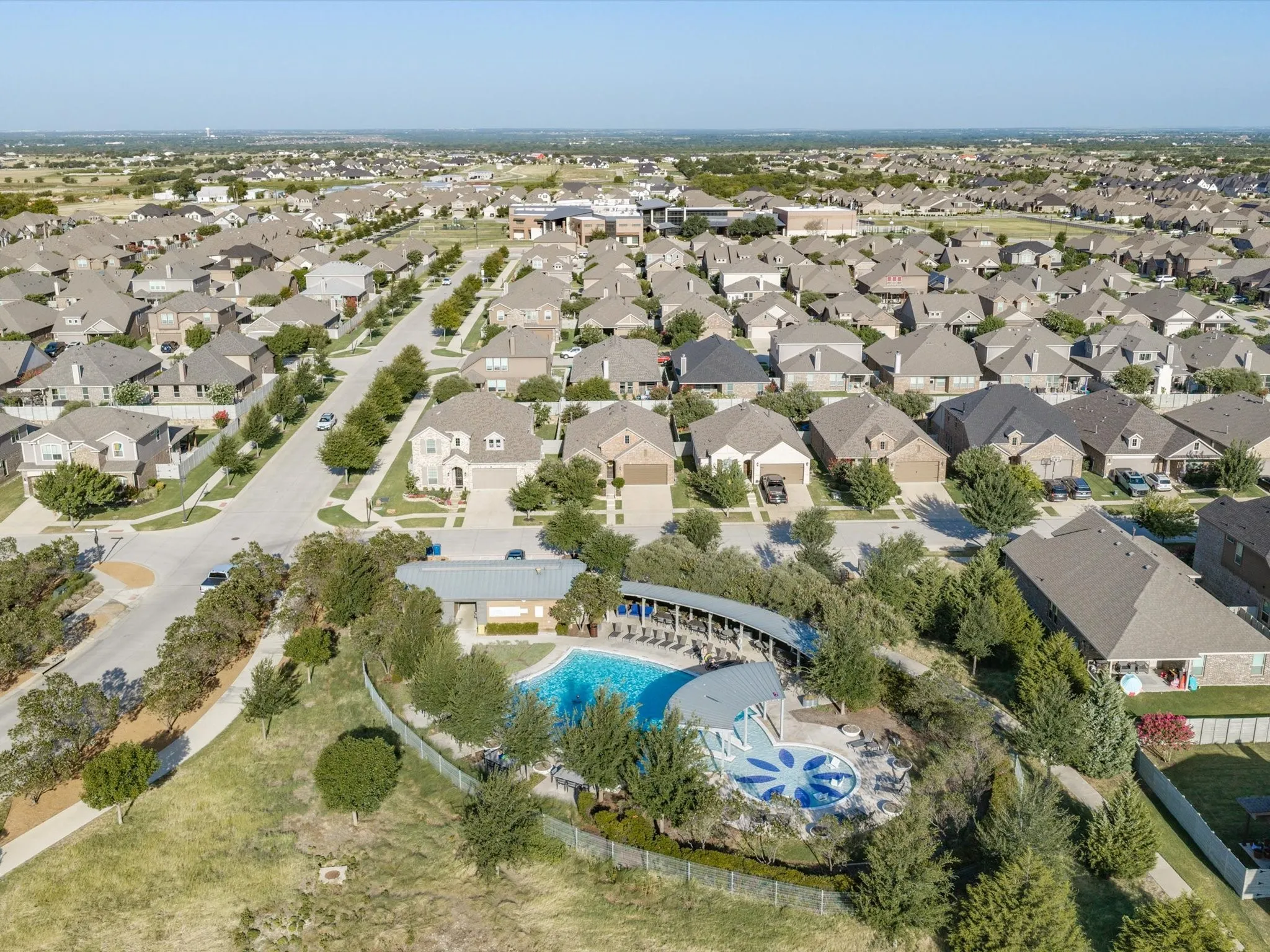 Aerial view showcasing a residential community with numerous single-family homes, a community pool with a sun shelf, and a covered amenity area