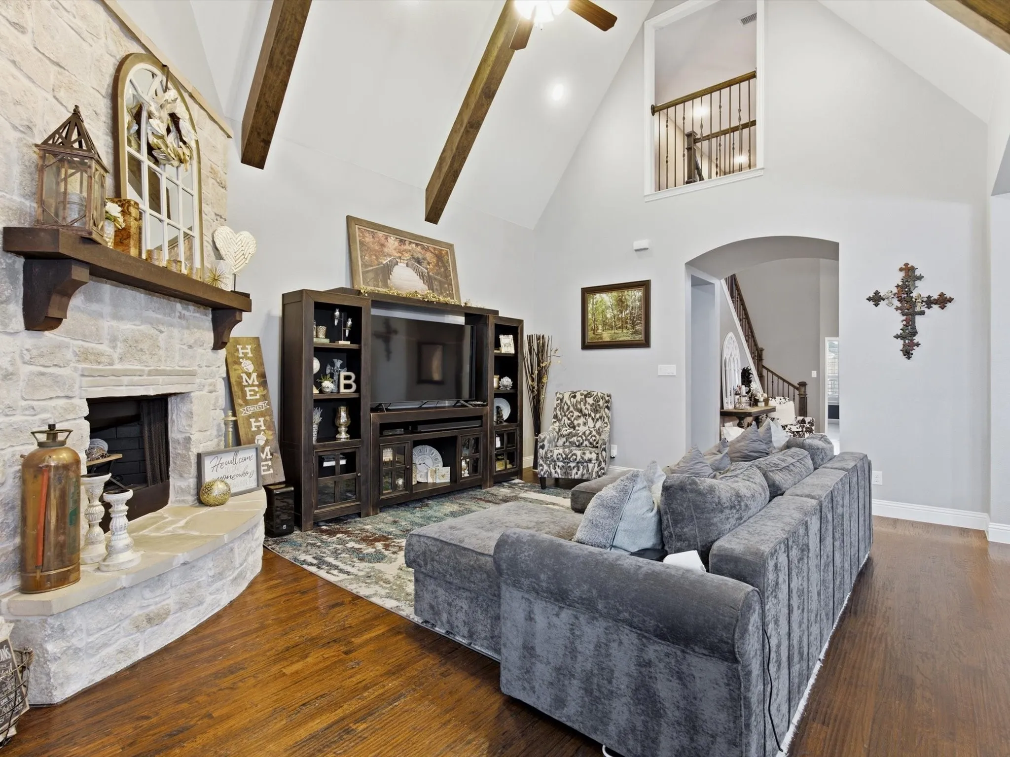 Living area featuring a stone-faced fireplace with a wood mantel, vaulted ceiling with exposed wood beams, wood-finish flooring, and a second-story overlook with a wrought iron railing