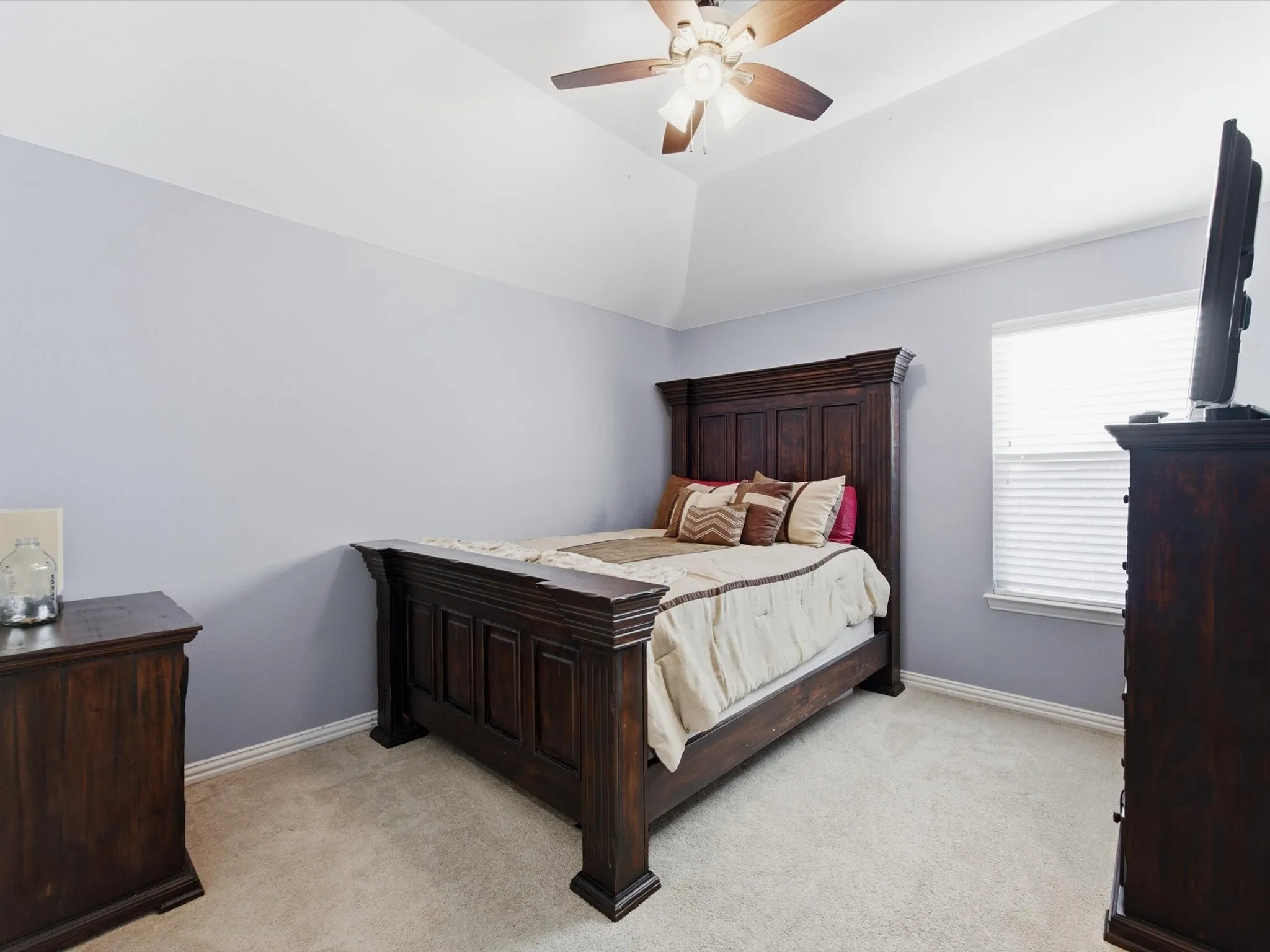 Vaulted ceiling bedroom with light gray walls, plush carpet flooring, a window with horizontal blinds, and a ceiling fan with light fixture
