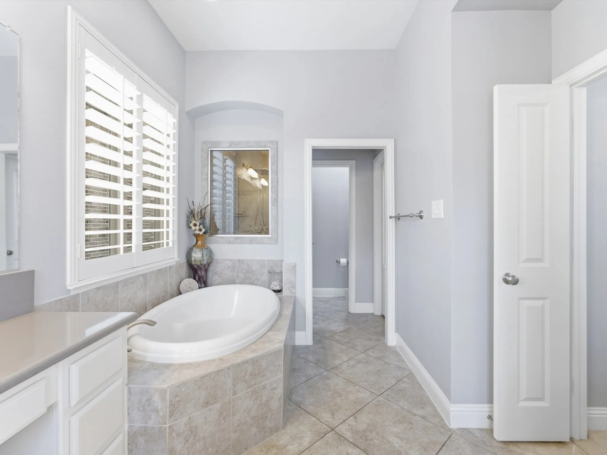 Soaking tub with tiled surround, plantation shutters, tiled flooring, and a vanity with built-in cabinetry and a light-toned countertop