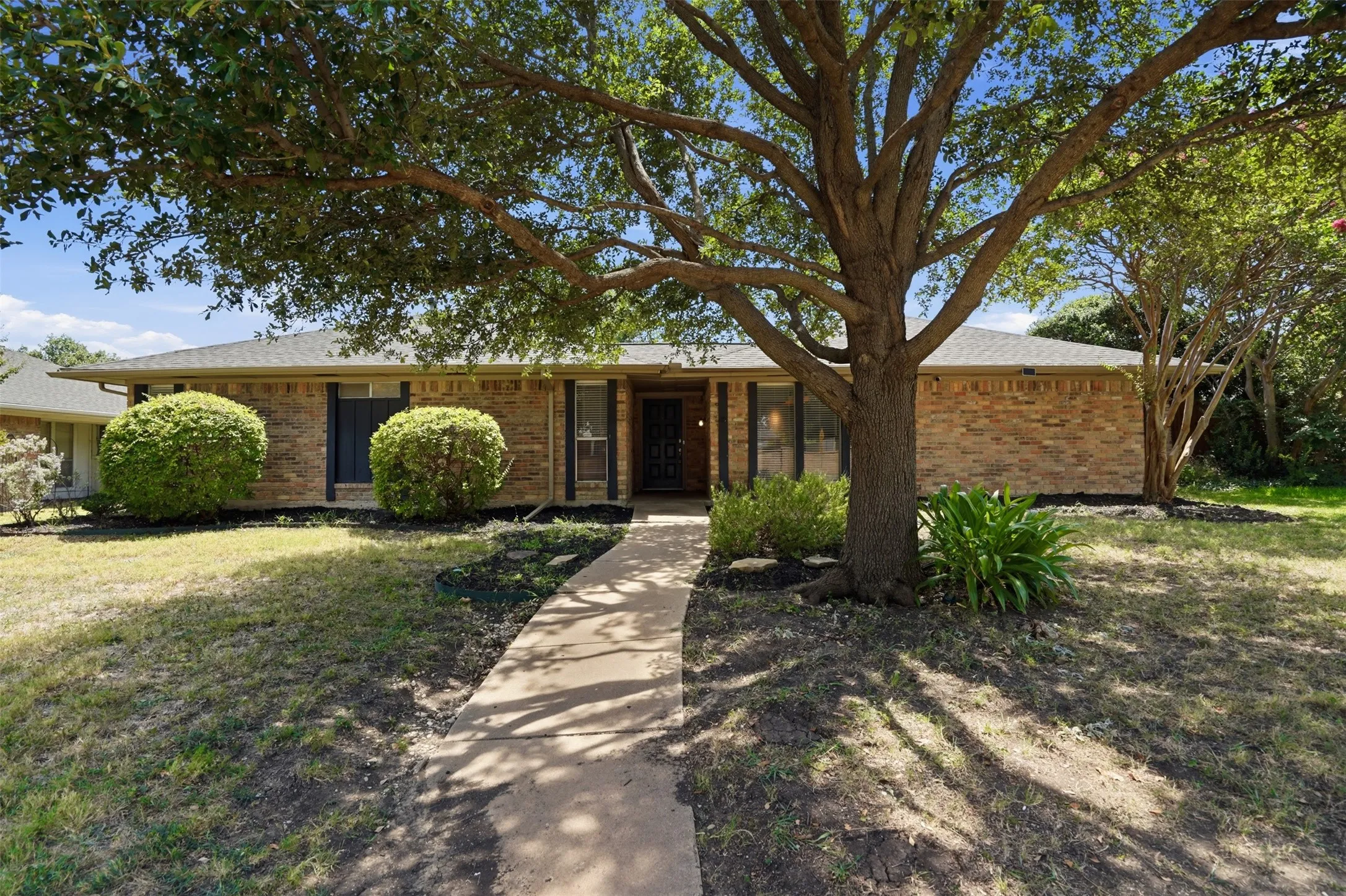 Brick exterior residence with a gray shingle roof, featuring a concrete pathway and mature trees providing shade