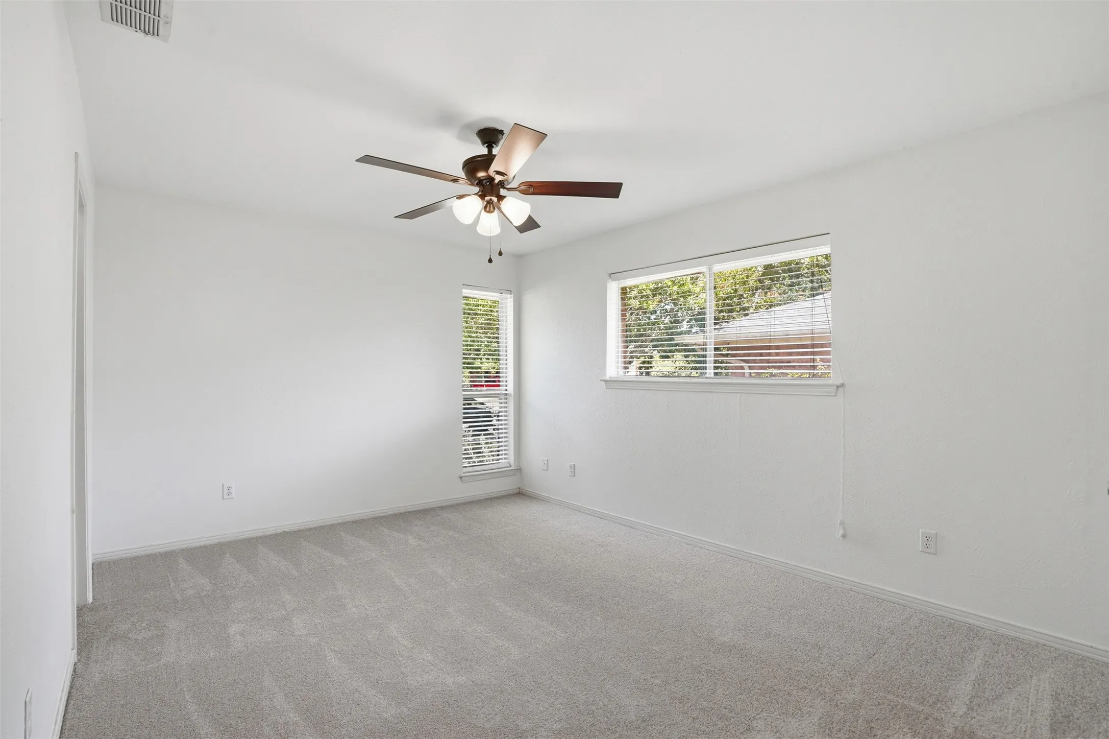 Spacious room with light gray carpeting, neutral white walls, a ceiling fan with integrated lighting, and two windows with blinds