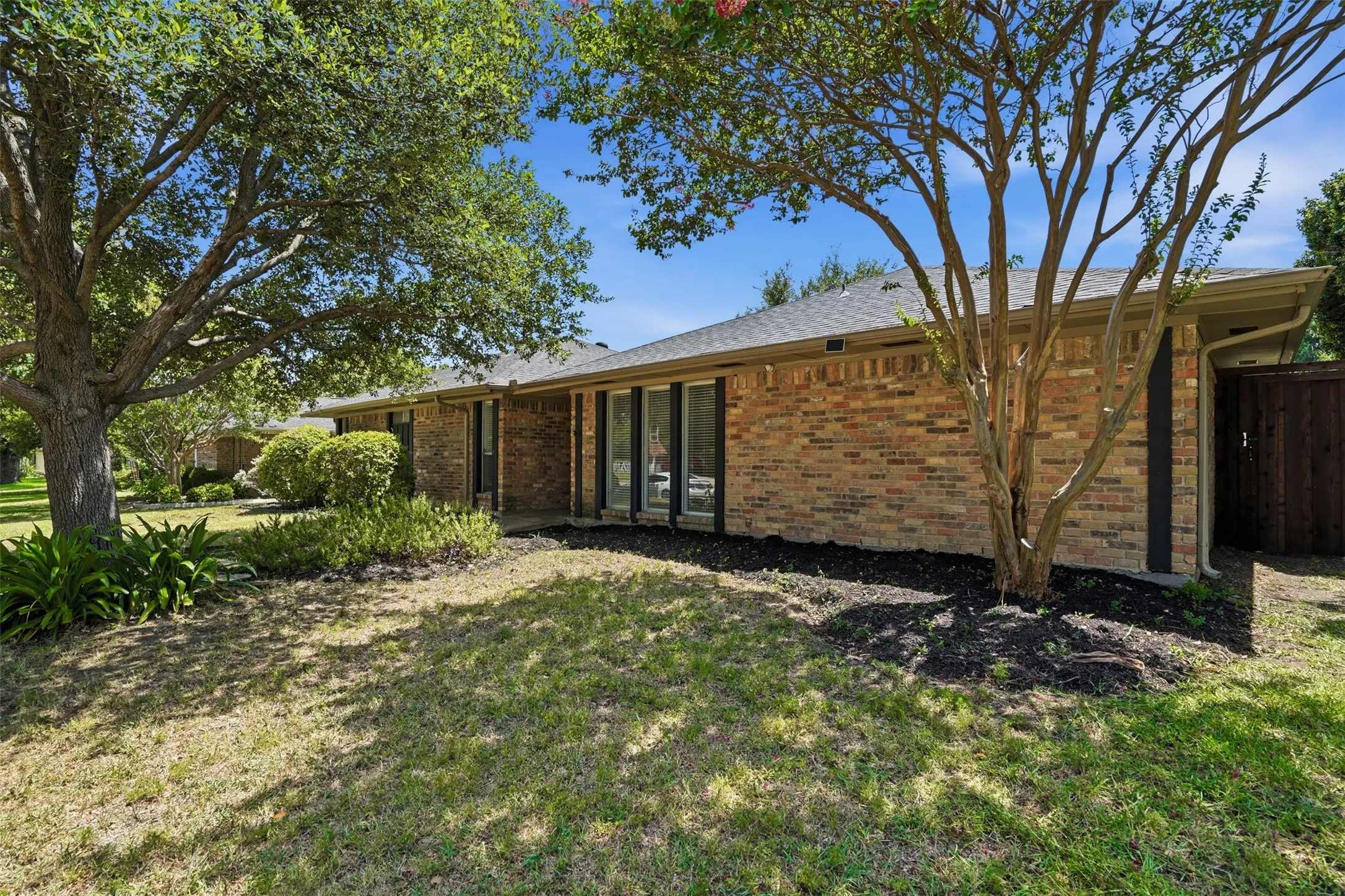 Brick exterior featuring a dark asphalt shingle roof, black-framed windows, mature trees, and established landscaping