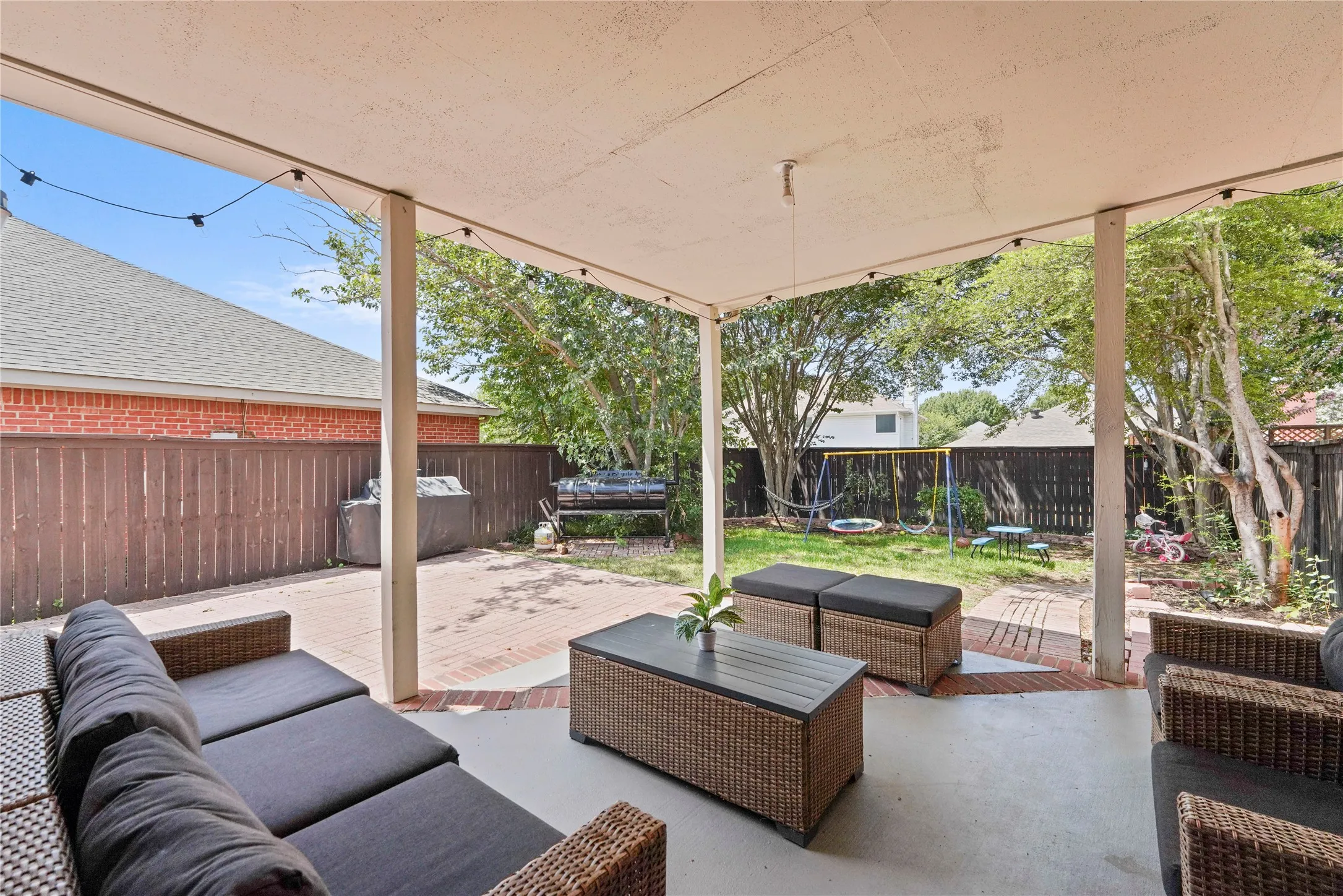 Covered patio featuring concrete and brick flooring, string lighting, and direct access to the private backyard with a wood fence perimeter