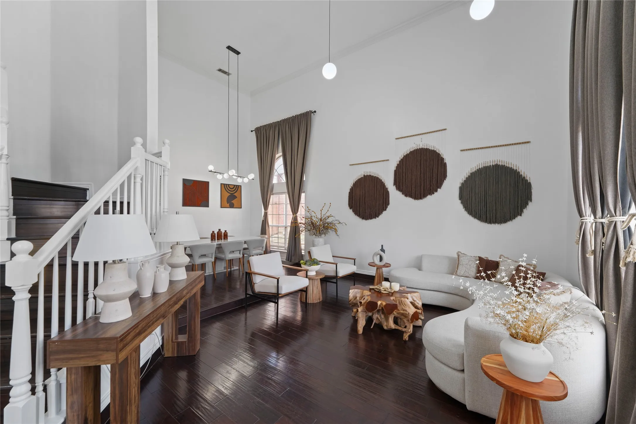 Spacious living area featuring dark wood-finish flooring, soaring ceilings, a white balustrade staircase, floor-to-ceiling drapery, and a modern linear chandelier