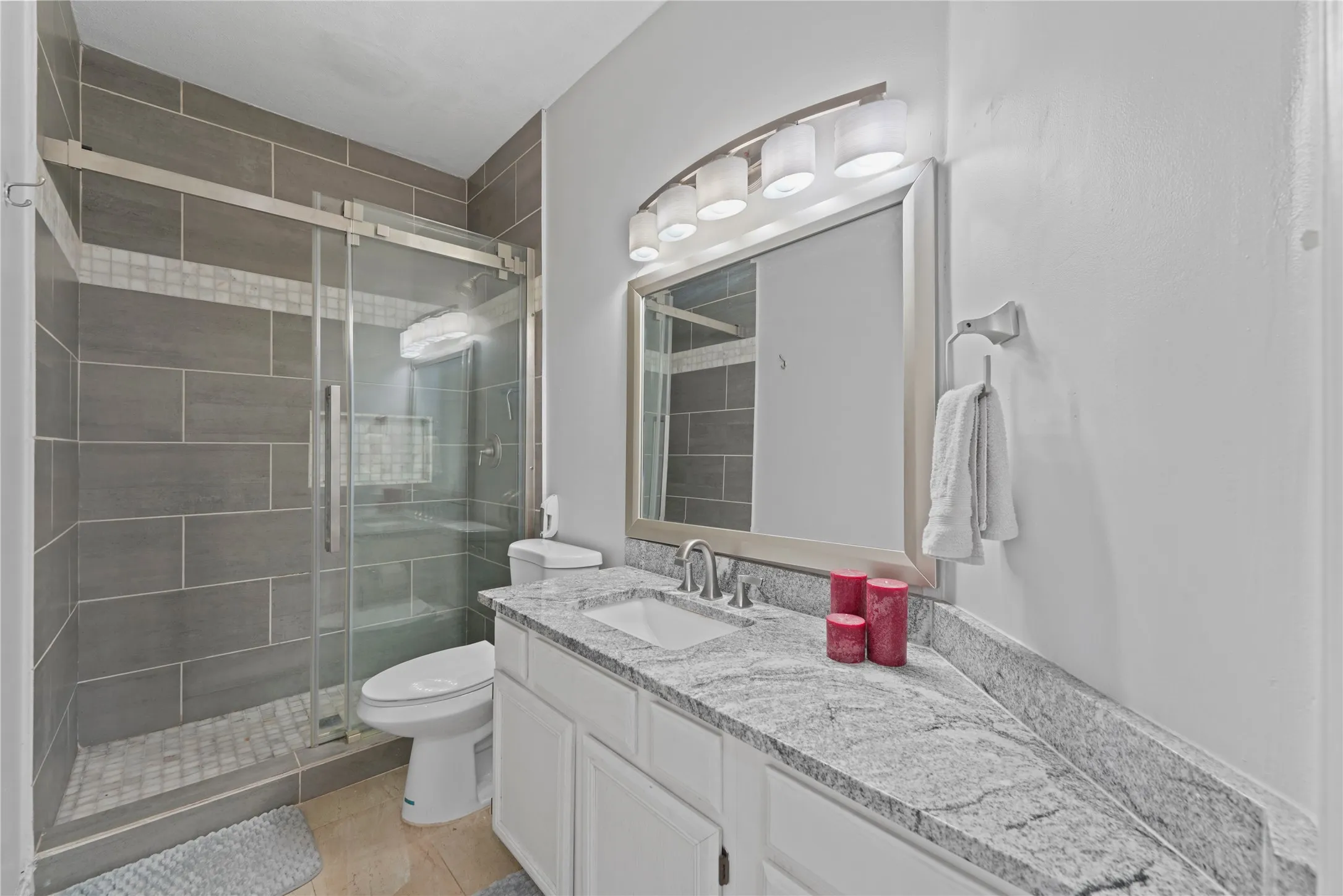 Bathroom featuring a glass-enclosed shower with gray subway tile surround and mosaic accent, white vanity with integrated sink, light-toned stone countertop, and silver-framed mirror with multi-bulb lighting fixture